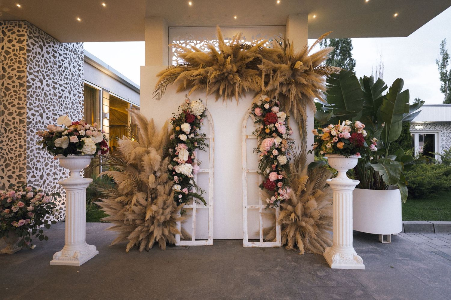 Elegant wedding arch with pampas grass and pink flowers on white pillars at an outdoor venue.