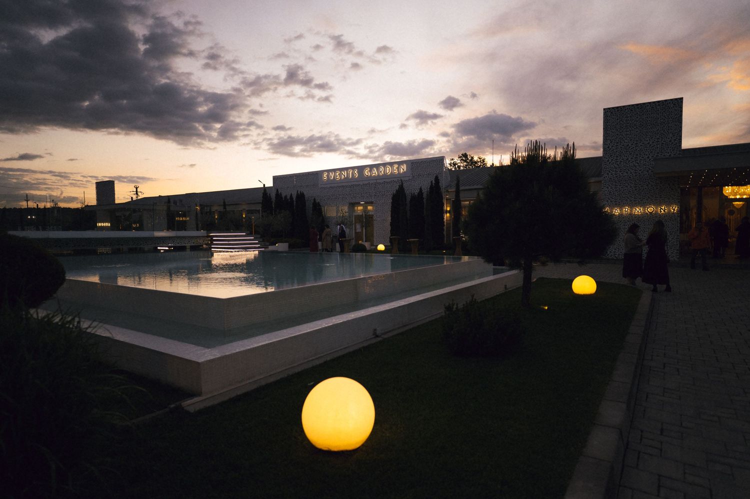 Illuminated globe lights glow at dusk beside a reflective pool at a modern outdoor venue.