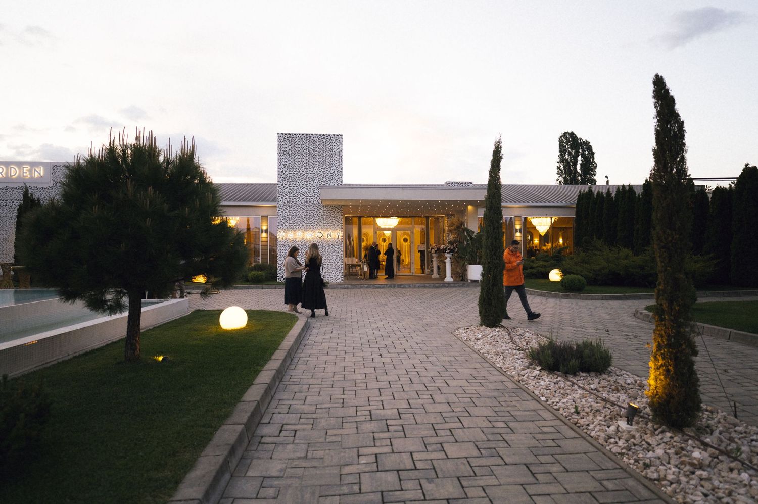 Elegant venue entrance at dusk with illuminated globe lights, cypress trees, and cobblestone path leading to a modern building.