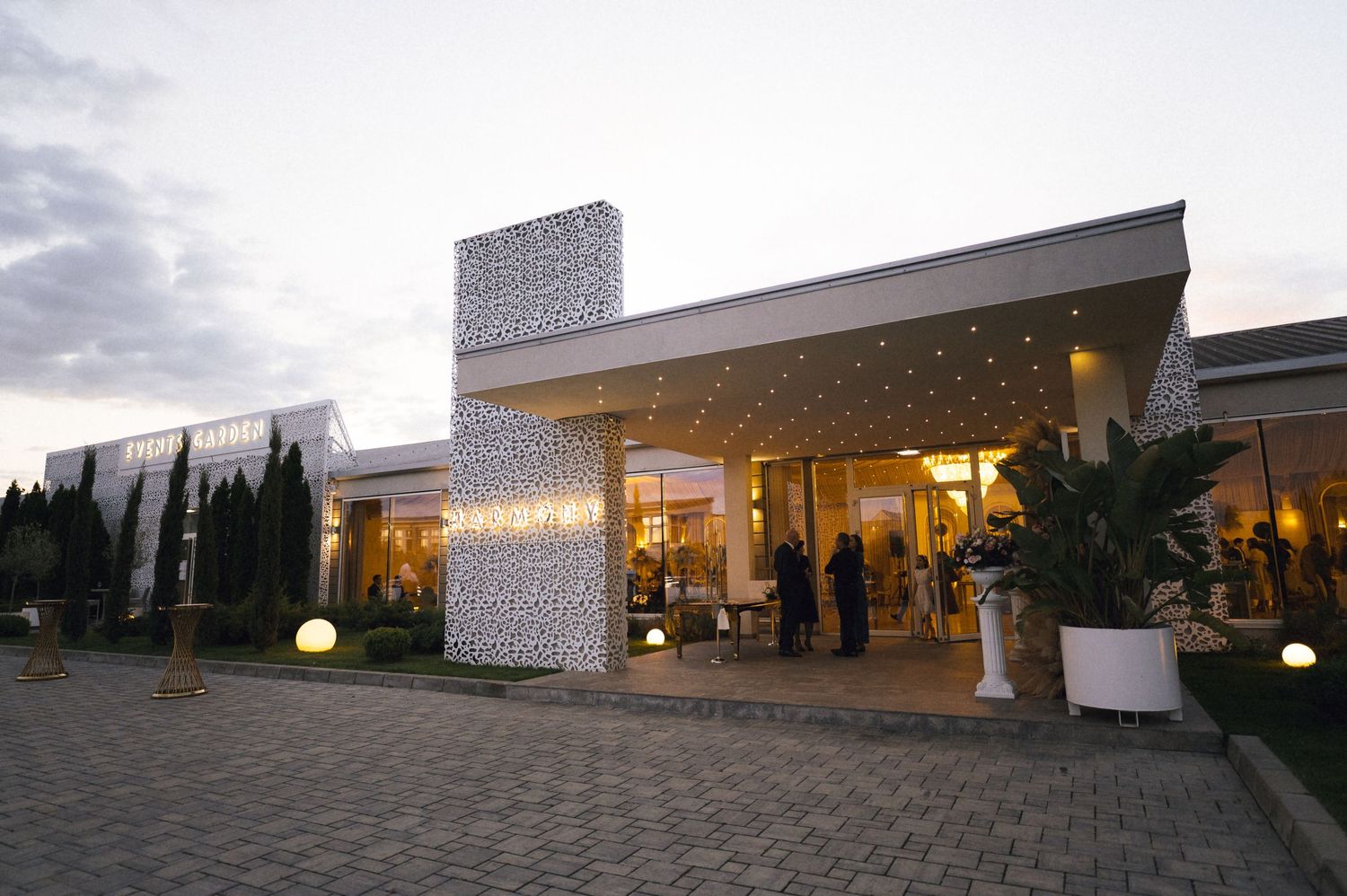 Elegant restaurant exterior illuminated at dusk with decorative white facade, lush greenery, and guests gathered at the entrance.