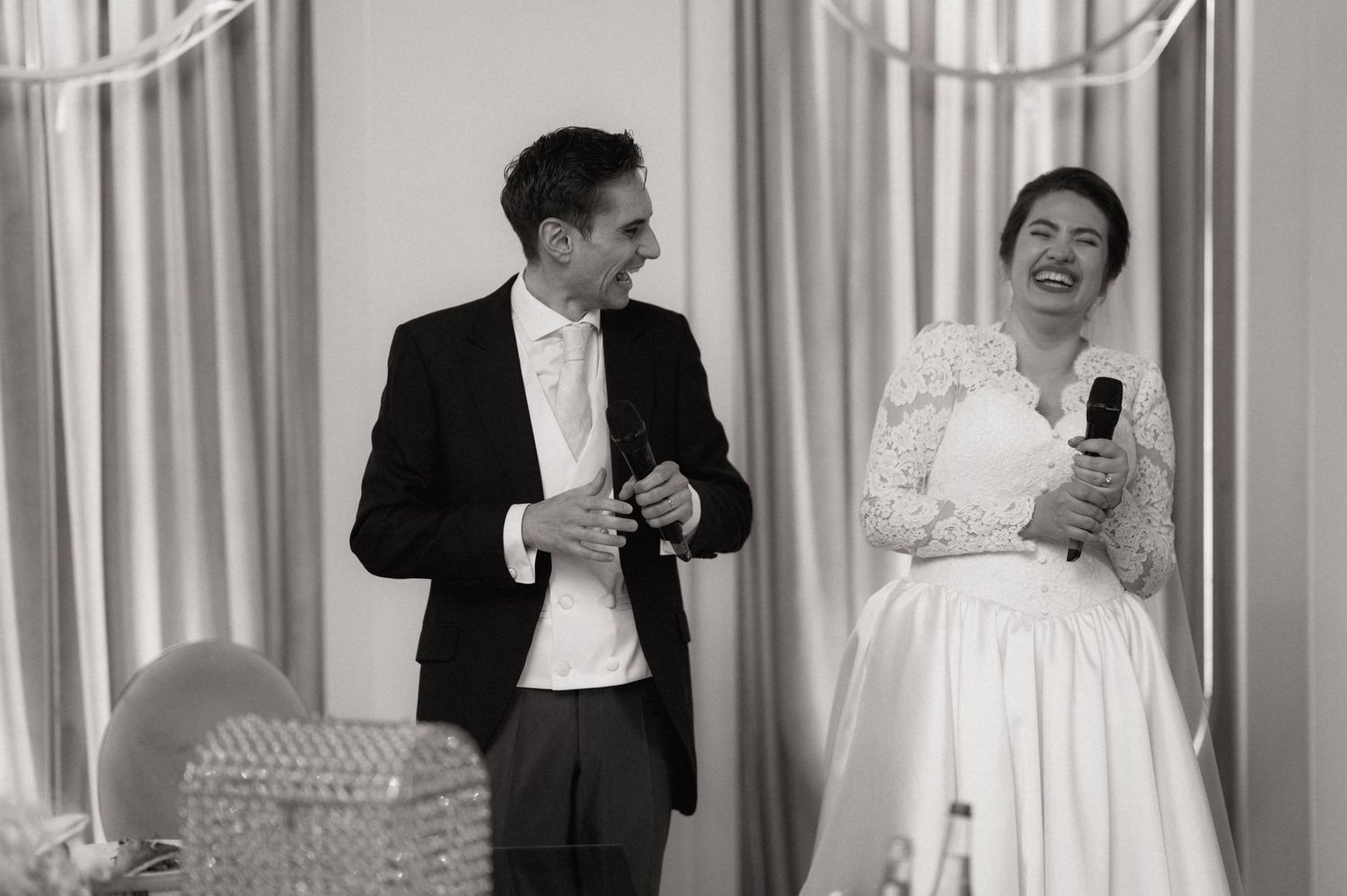 Black and white photo of a laughing bride in lace dress and groom in tuxedo at wedding reception.