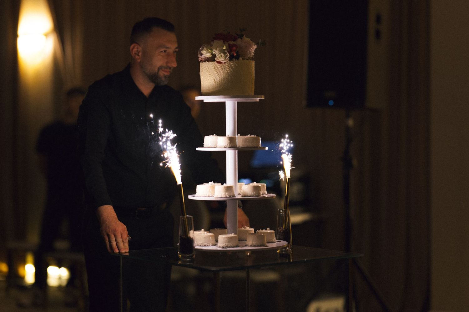 Man in black stands beside a elegant multi-tier wedding cake display with sparkling fireworks at a dark reception.