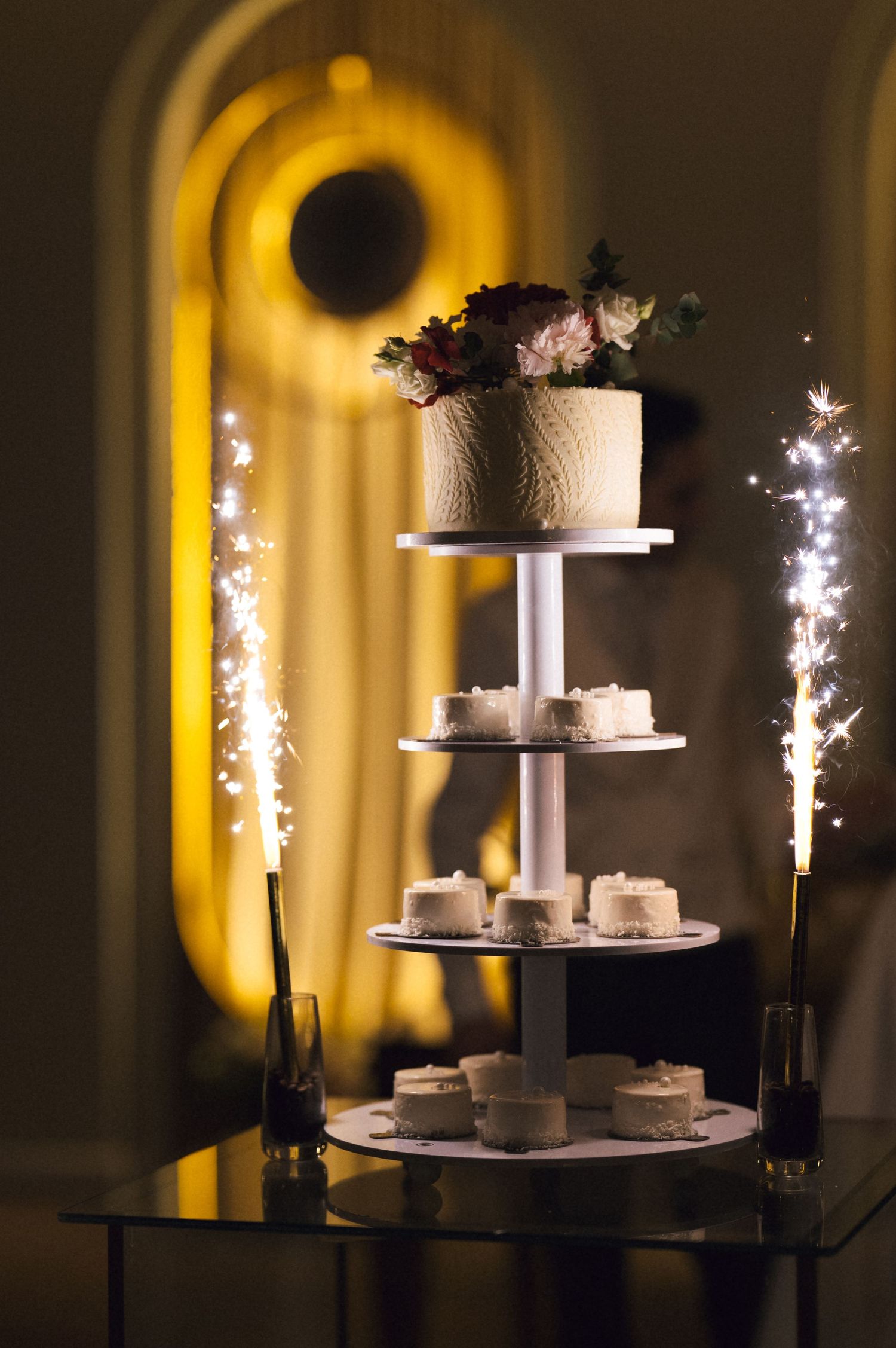 Multi-tier wedding cake with sparklers and floral topper displayed on a table in a warmly lit elegant venue.