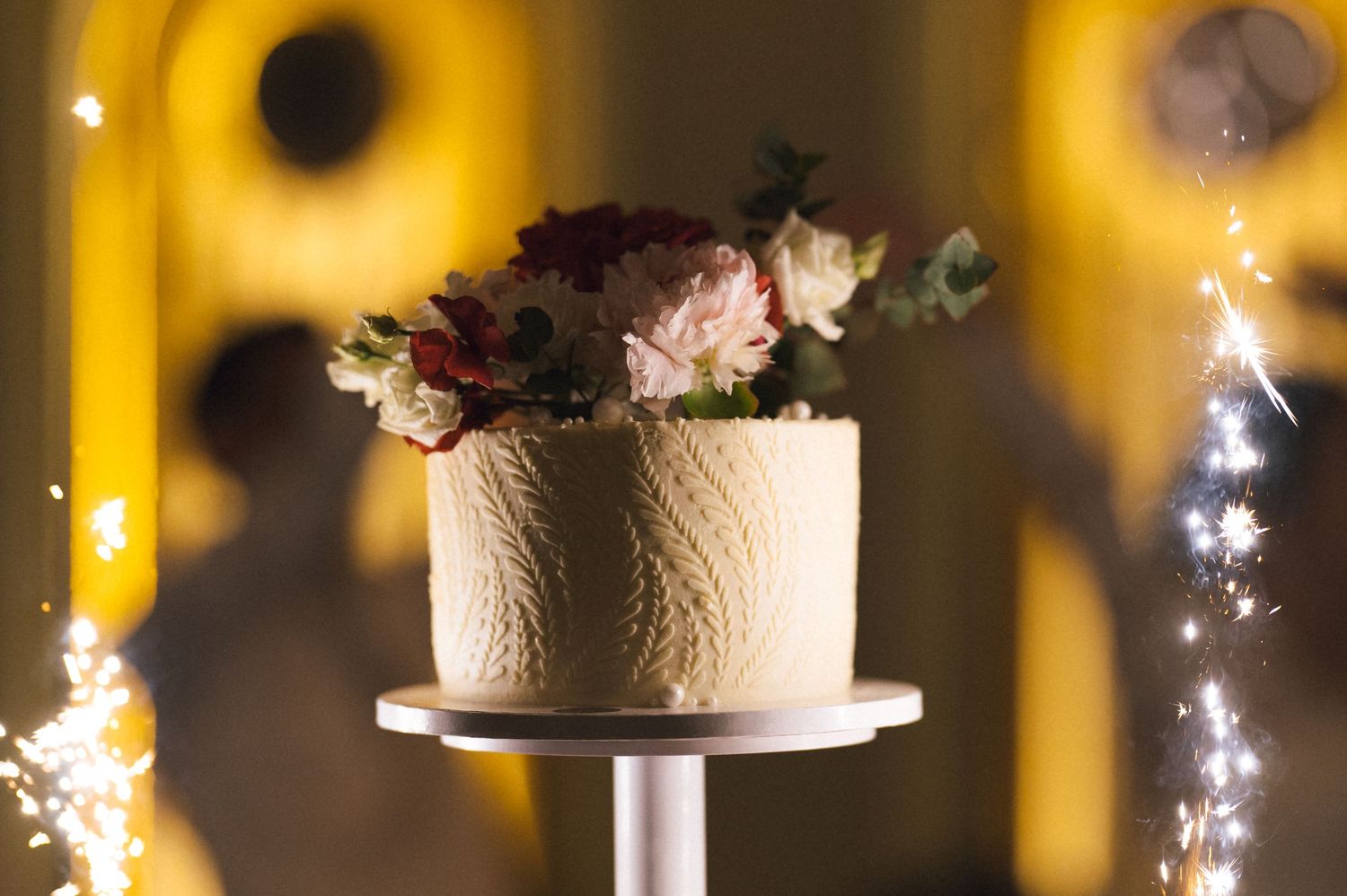 Elegant white textured wedding cake topped with fresh flowers on a pedestal stand, surrounded by sparkling fairy lights.