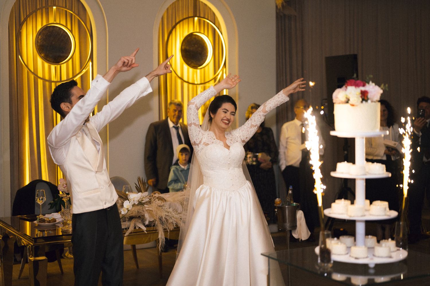 Bride and groom dancing joyfully at wedding reception with sparklers and elegant white tiered cake in background.