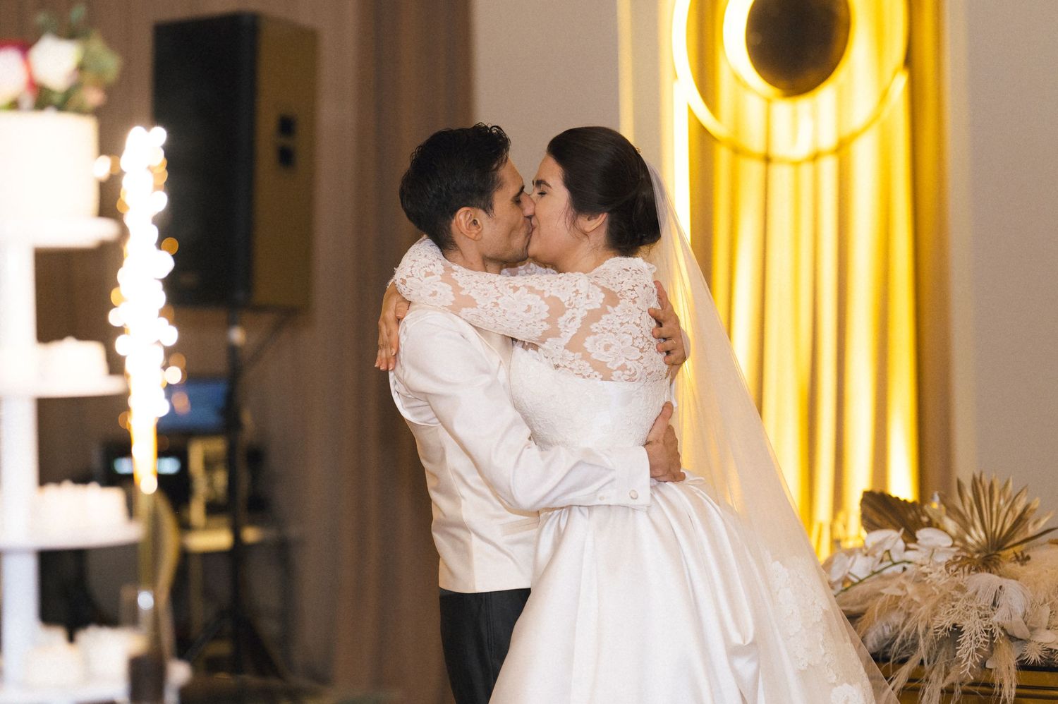 A bride and groom share a romantic first kiss at their elegant wedding reception with warm golden lighting.
