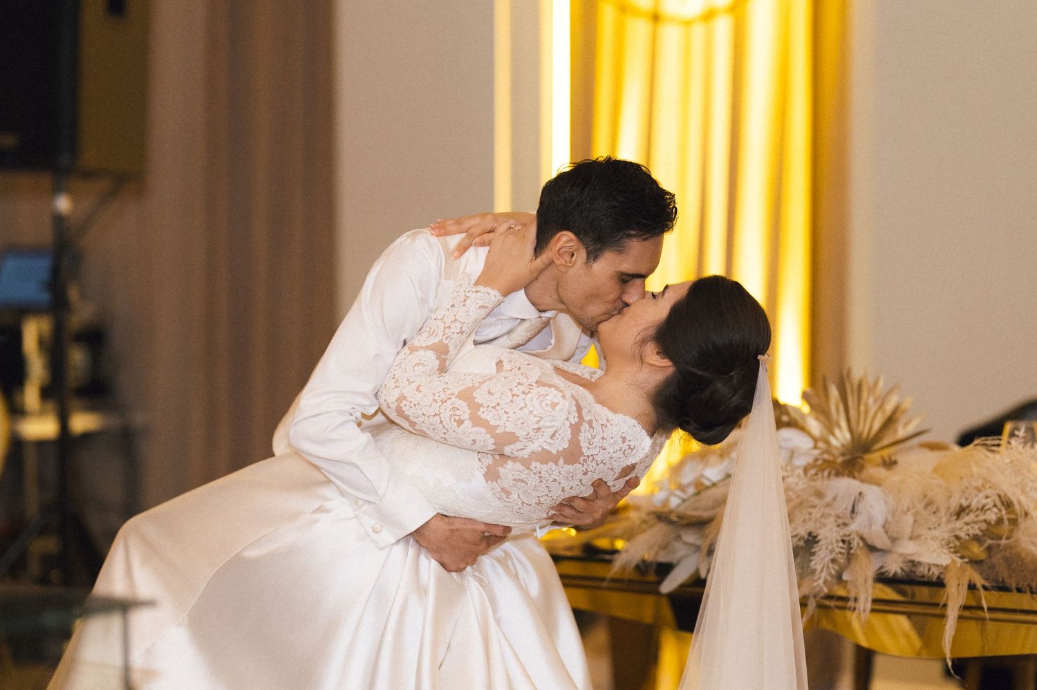 Bride and groom share a romantic dip kiss at their wedding reception, with golden curtains and warm lighting in the background.
