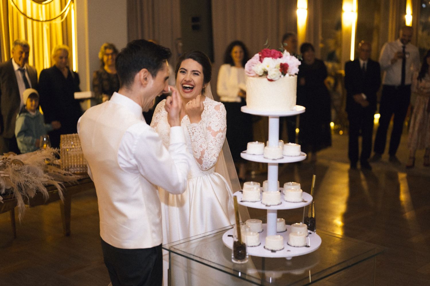 Bride and groom laughing while cutting their elegant white wedding cake at a romantic indoor reception.