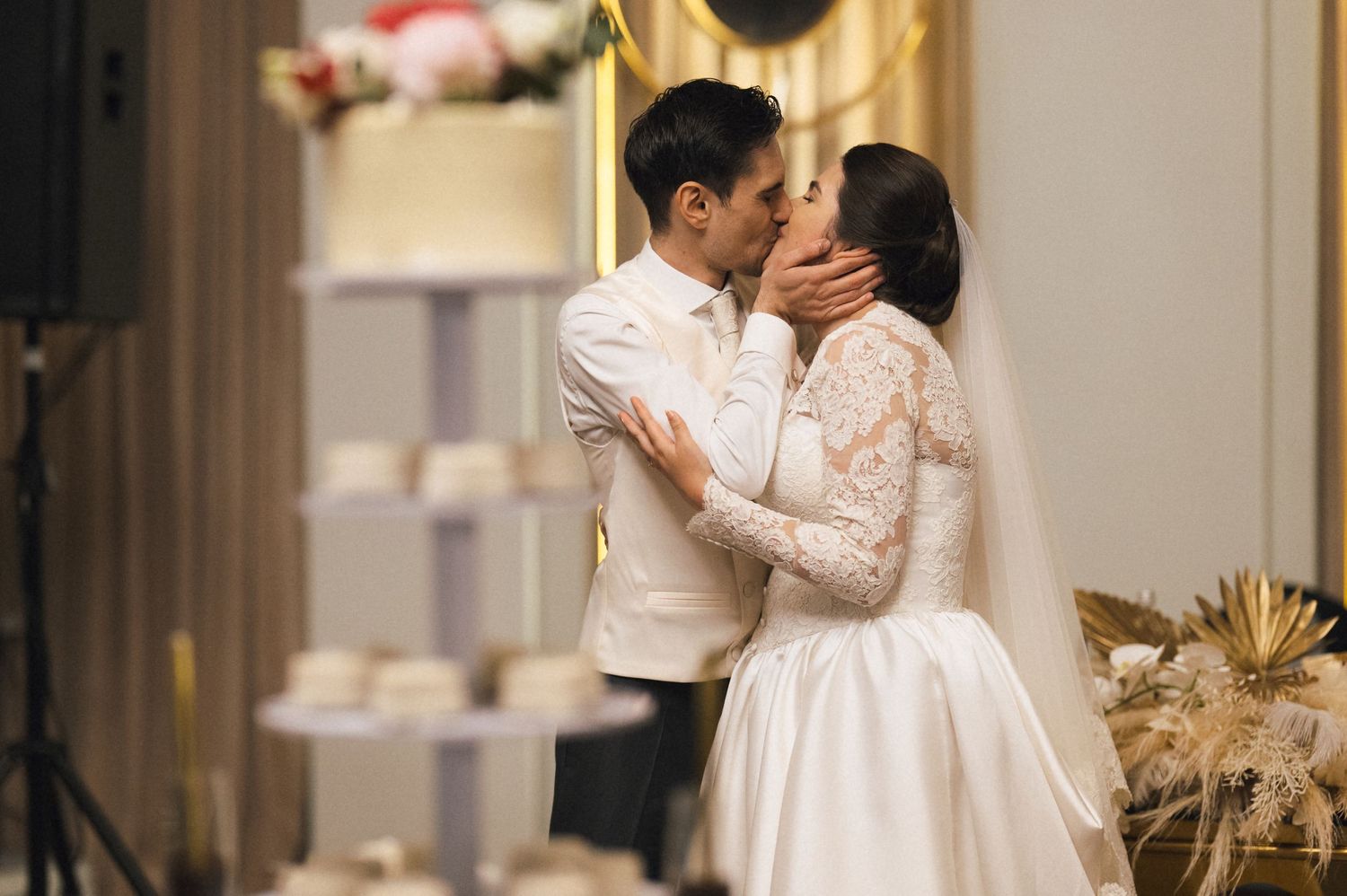 Bride and groom share a romantic first kiss at their elegant wedding reception near a tiered white cake.