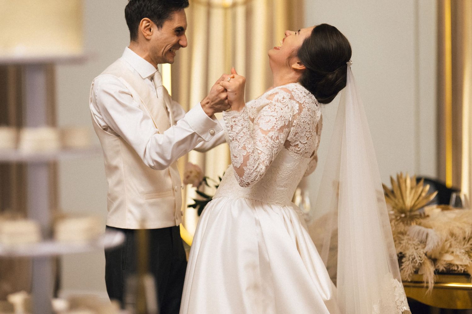 A bride and groom share their first dance in an elegant venue, the bride wearing a lace long-sleeve wedding gown with veil.