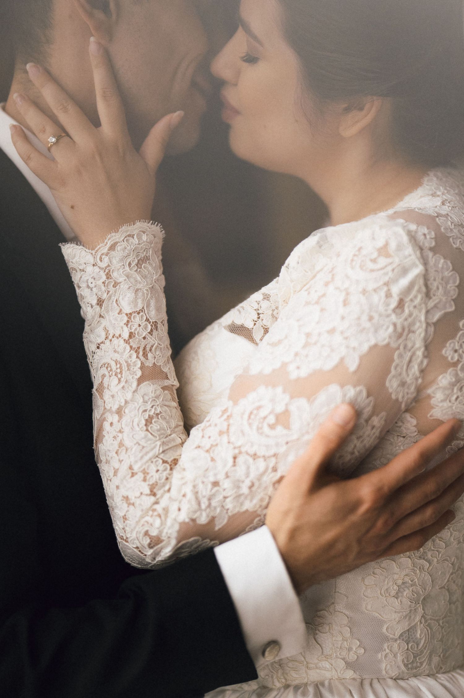 Bride in long-sleeve lace wedding gown embraces groom in black suit as they share a romantic kiss.