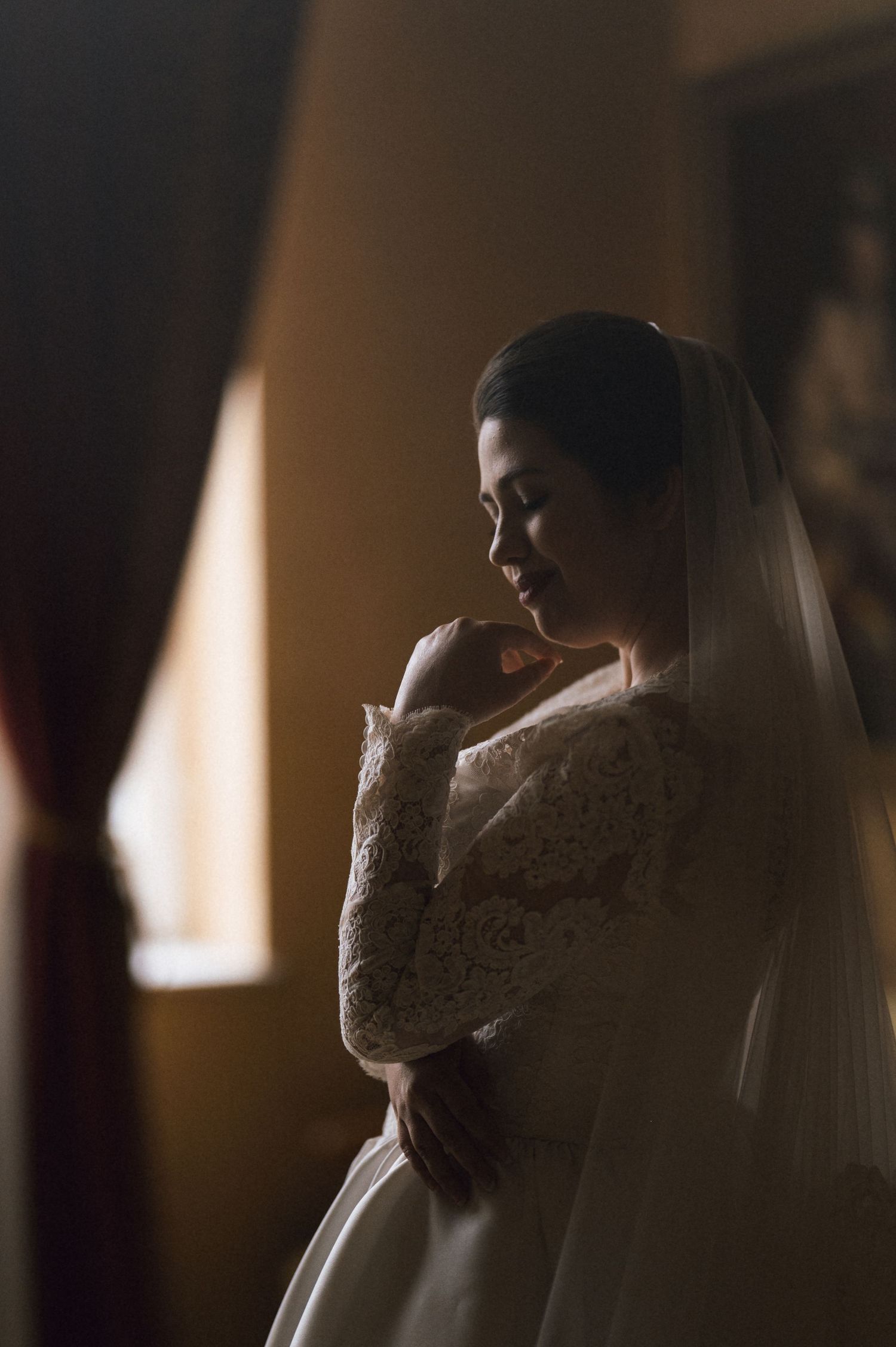 Bride in lace long-sleeve wedding gown and veil stands by window in soft golden light, eyes closed in serene moment.
