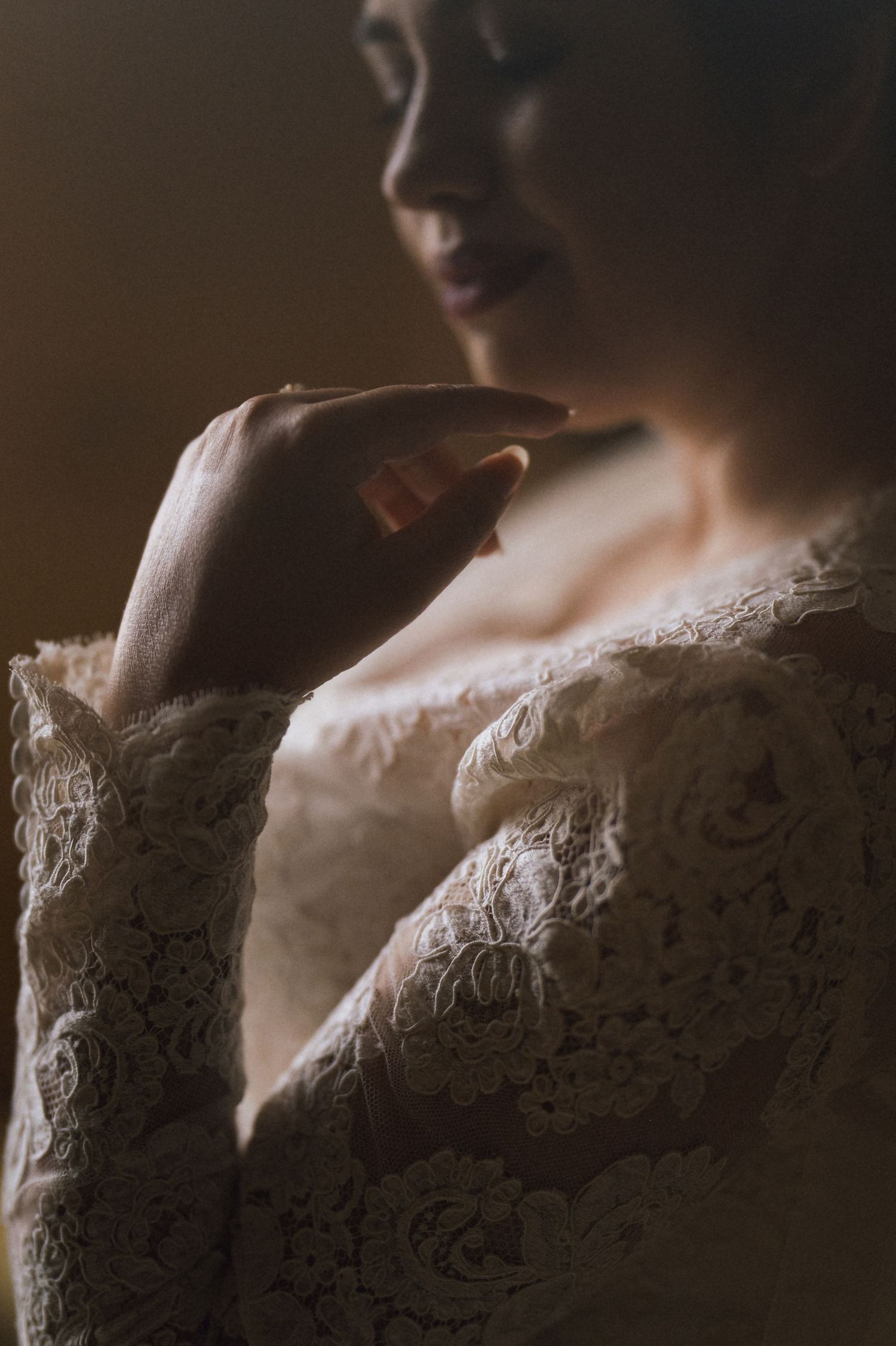 A bride in a delicate white lace long-sleeve gown gently touches her shoulder in a moody, softly lit portrait.