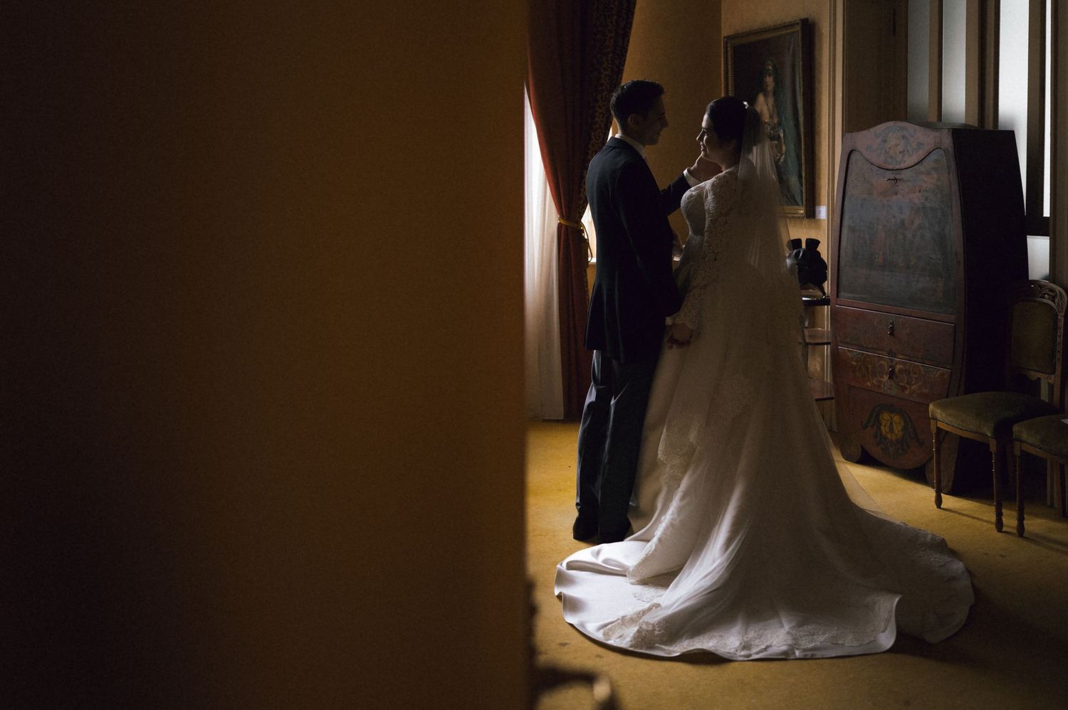 Bride in elegant white gown with long train stands with groom in dark suit in dimly lit room.