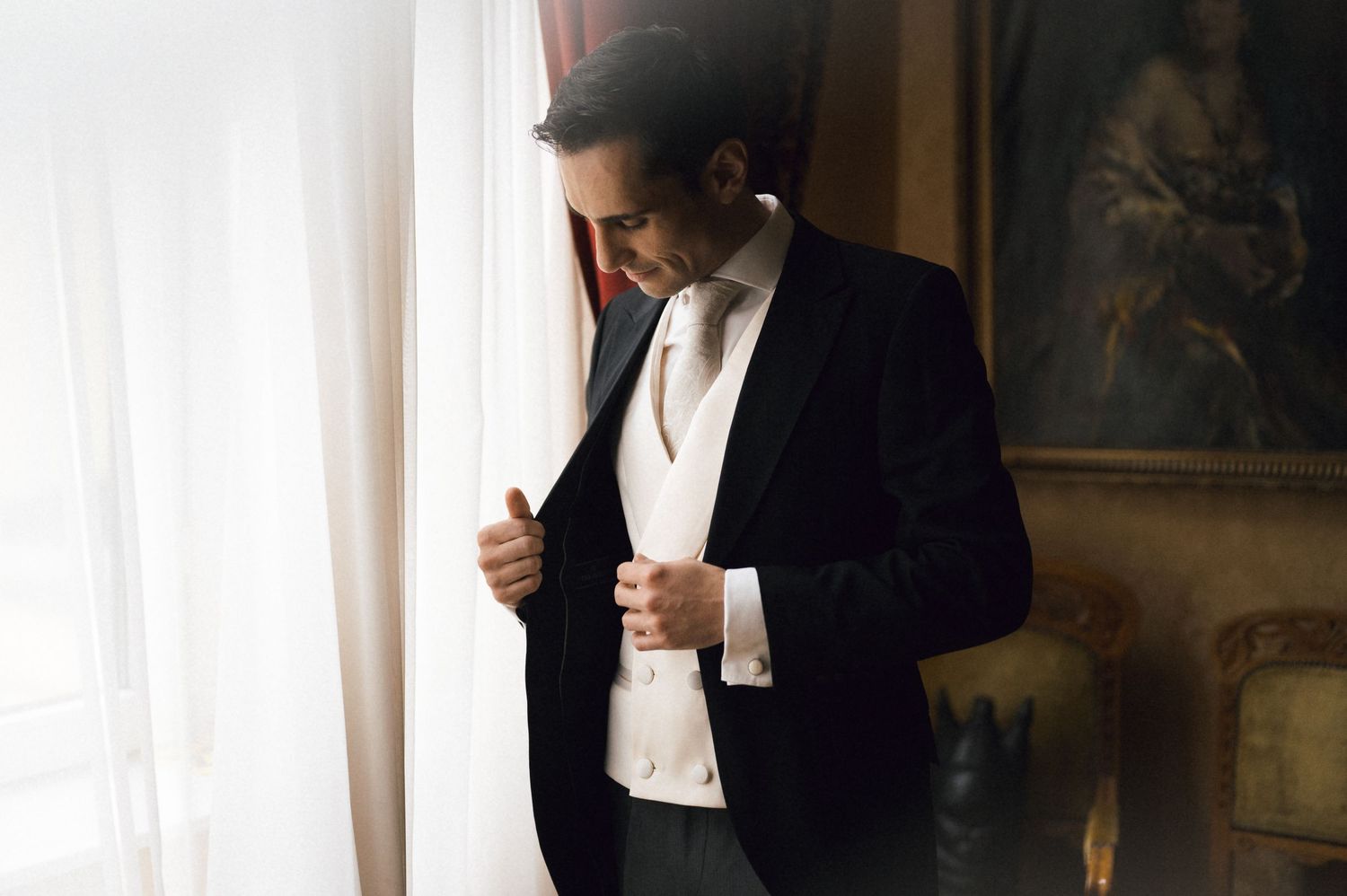 Elegant groom in a dark suit and white vest buttoning his jacket near a window in a classic interior.