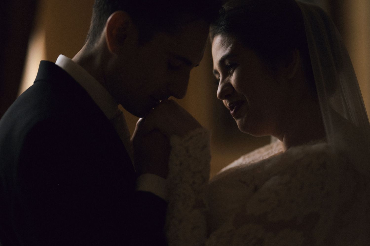 Romantic wedding couple in elegant attire sharing an intimate moment in soft dramatic lighting.