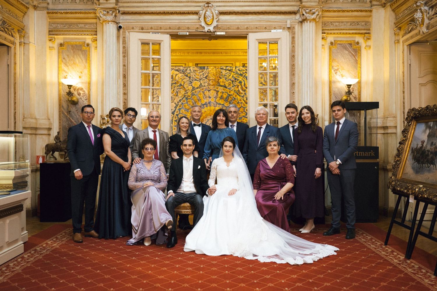 Wedding party group photo with bride in white gown surrounded by family in formal attire in ornate room.