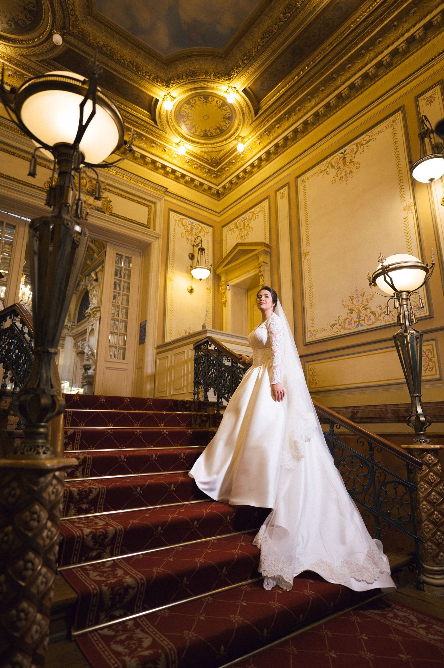 Bride in elegant white gown with long train stands on ornate red-carpeted staircase in a grand golden ballroom.