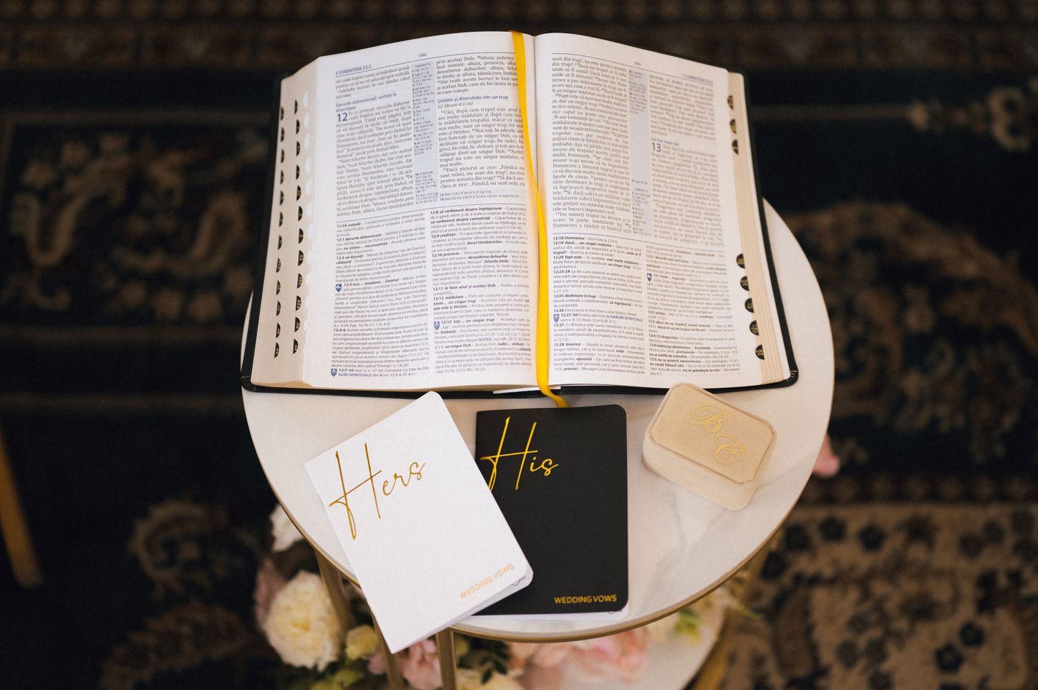 Open Bible with yellow bookmark beside His and Hers journals on a white table with flowers.