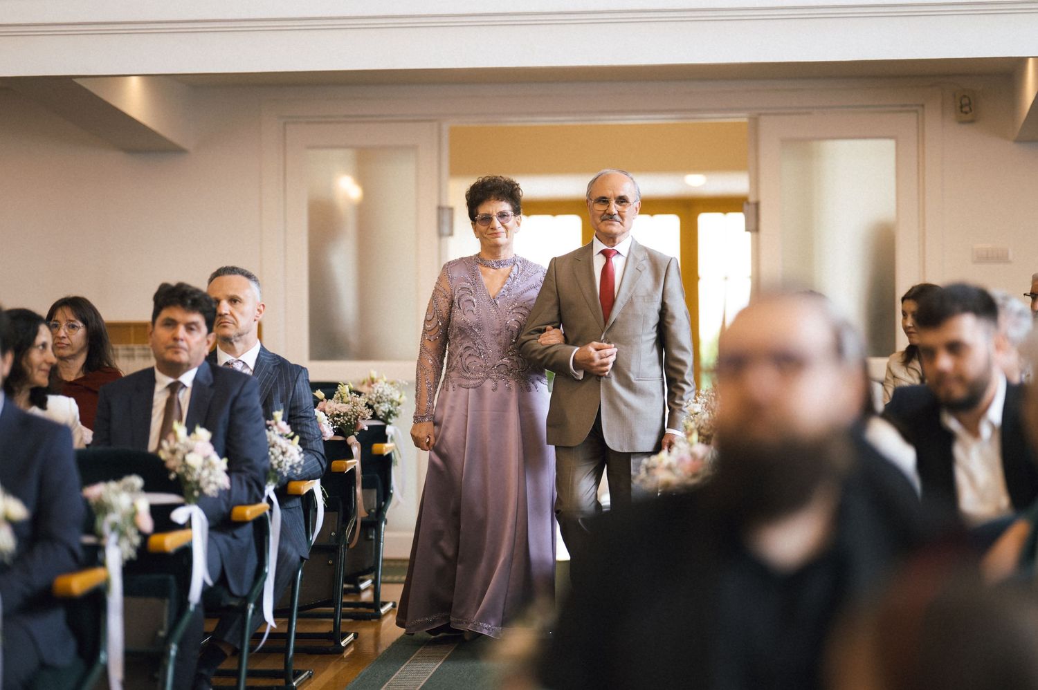 Elegant couple walks down wedding aisle together, surrounded by seated guests in a formal venue.