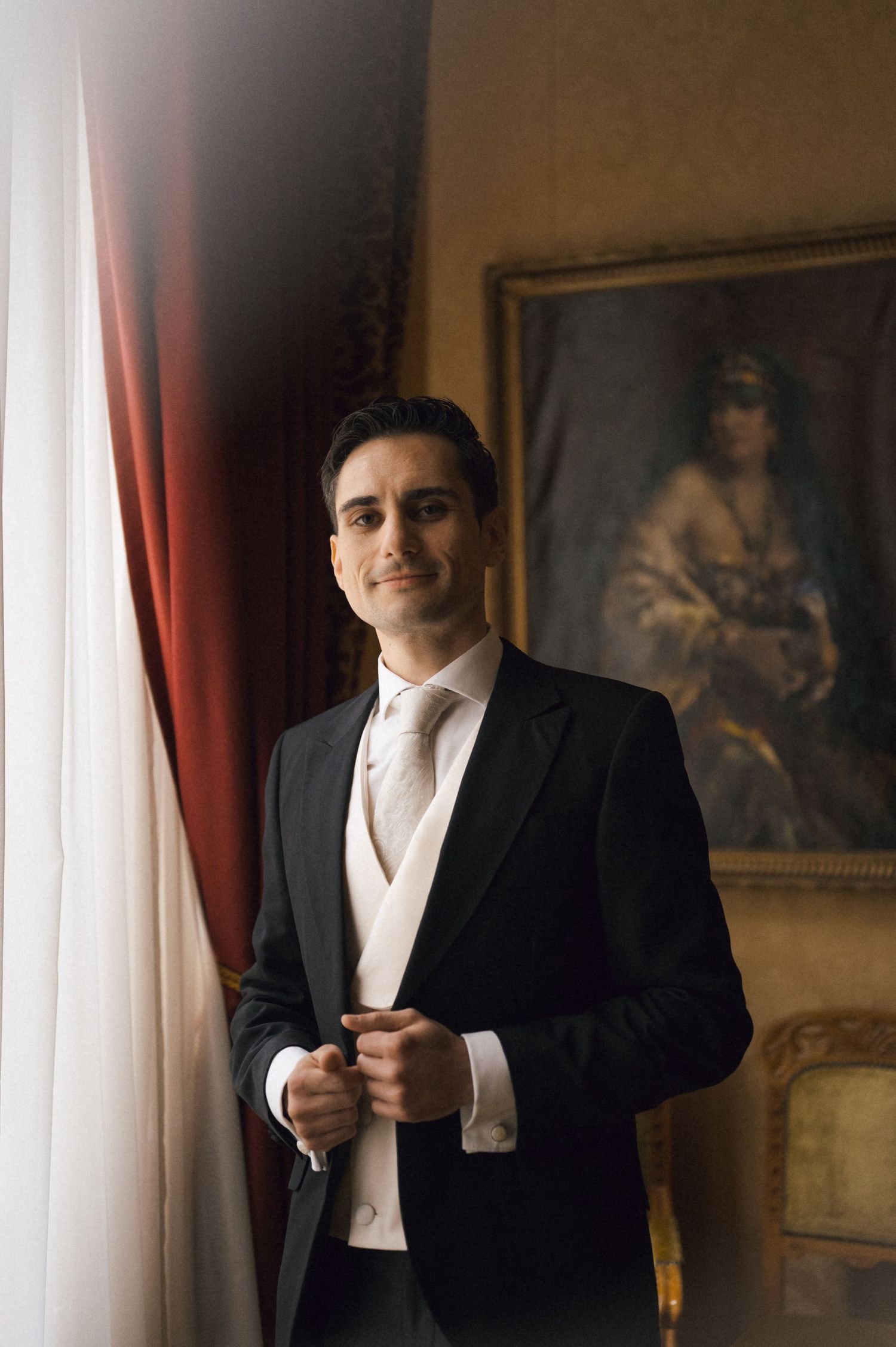 Elegant groom in black tuxedo and white vest adjusting jacket near window with red curtains and classic portrait painting.