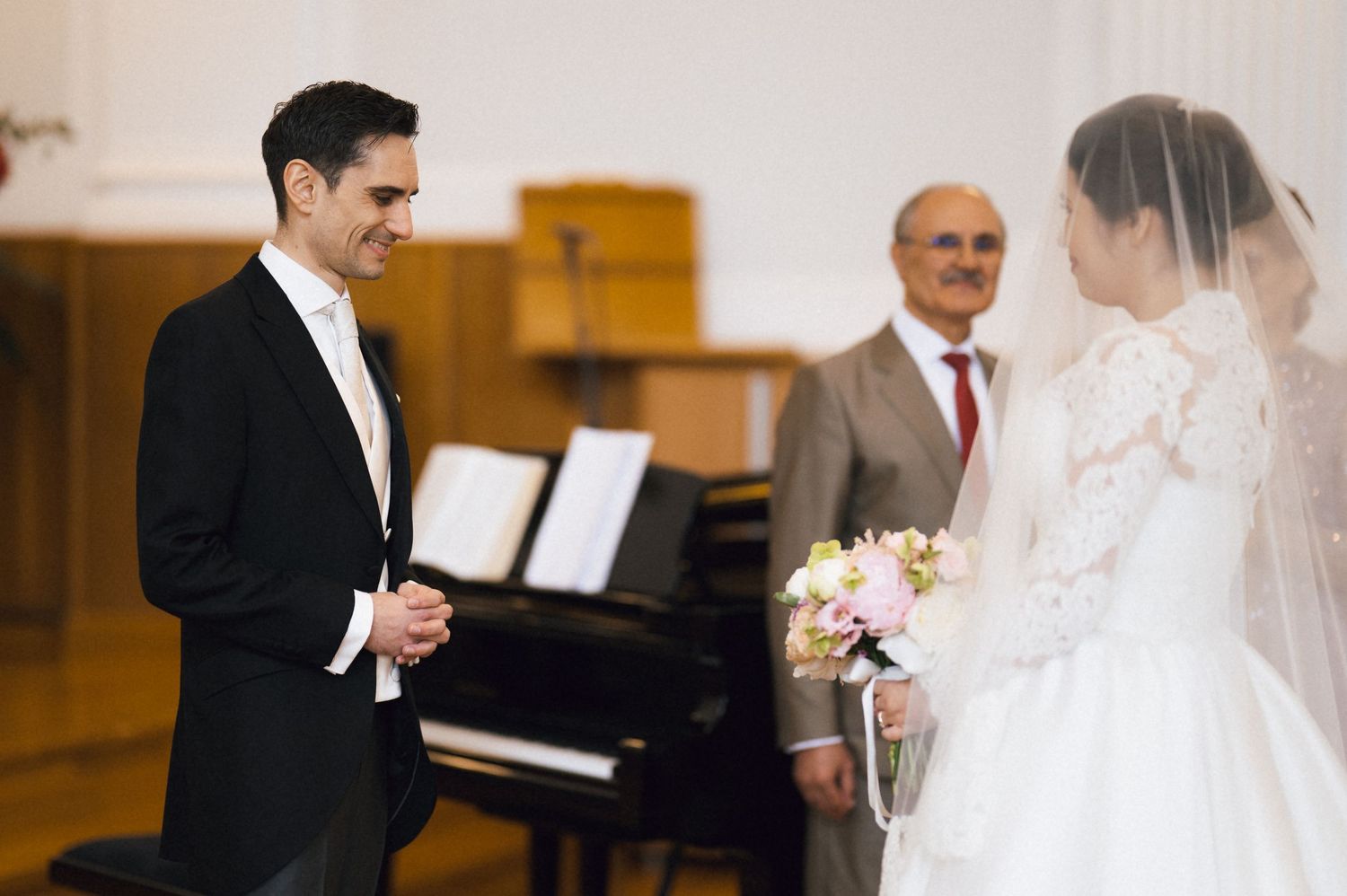 Groom in black tuxedo smiles at bride in white lace dress holding bouquet during church wedding ceremony.