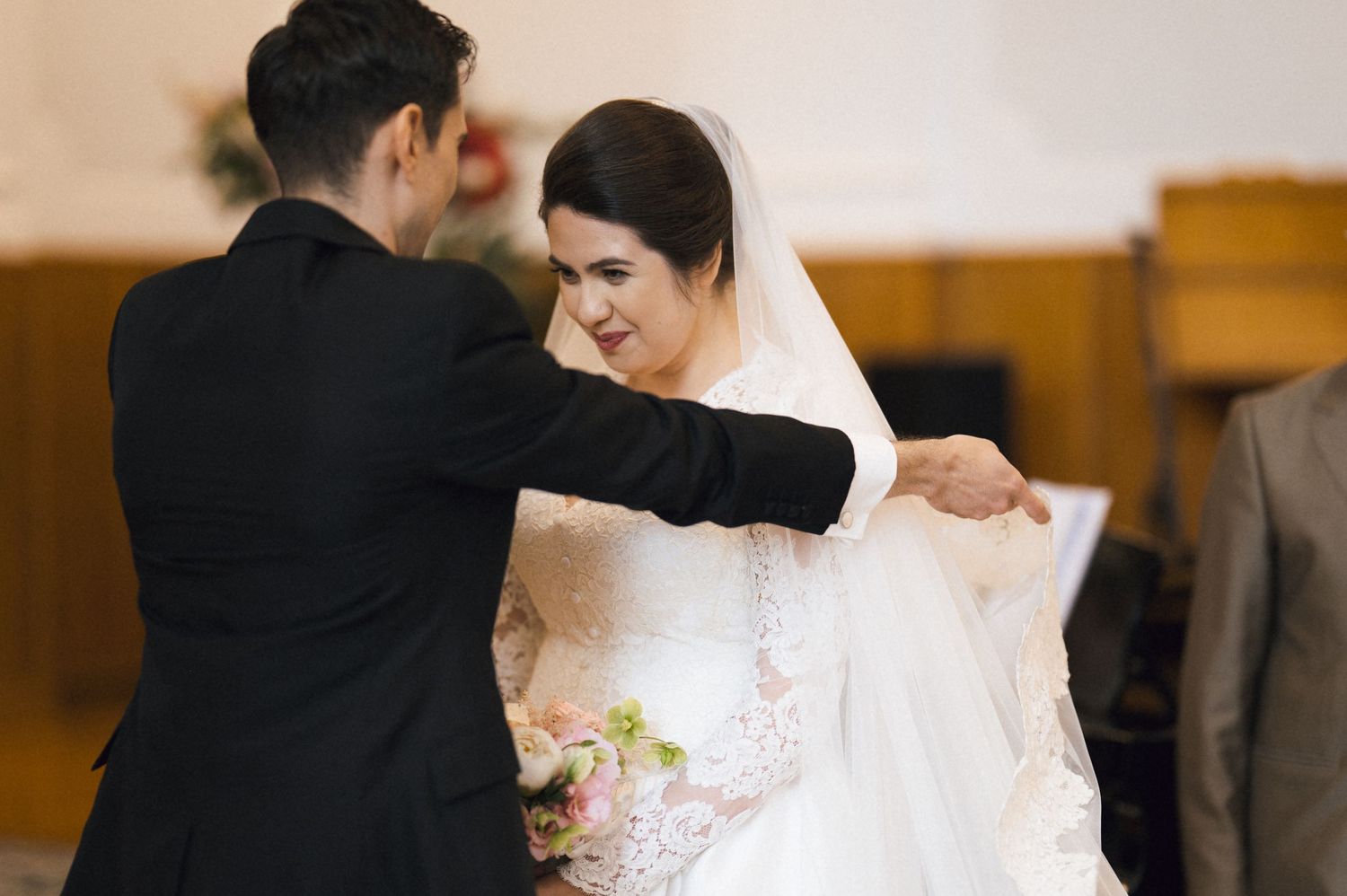 Groom in black suit adjusting bride's veil during wedding ceremony in church, bride holding bouquet.
