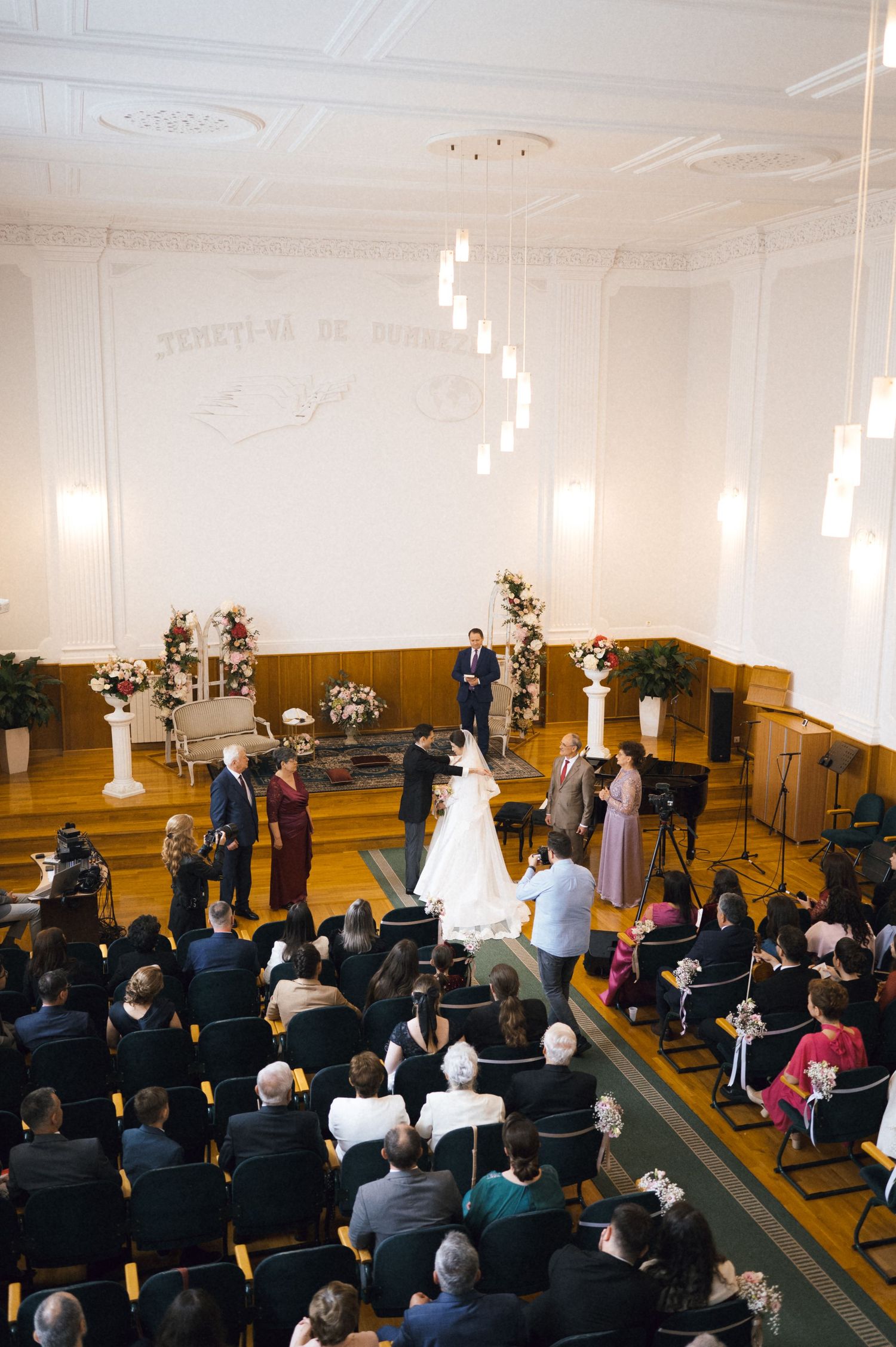 Elegant wedding ceremony in a grand hall with guests seated, bride in white gown at the altar with floral arrangements.