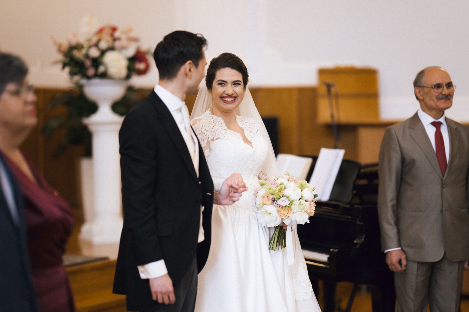 Bride and groom smiling during wedding ceremony in church, bride holding white bouquet, guests visible in background.