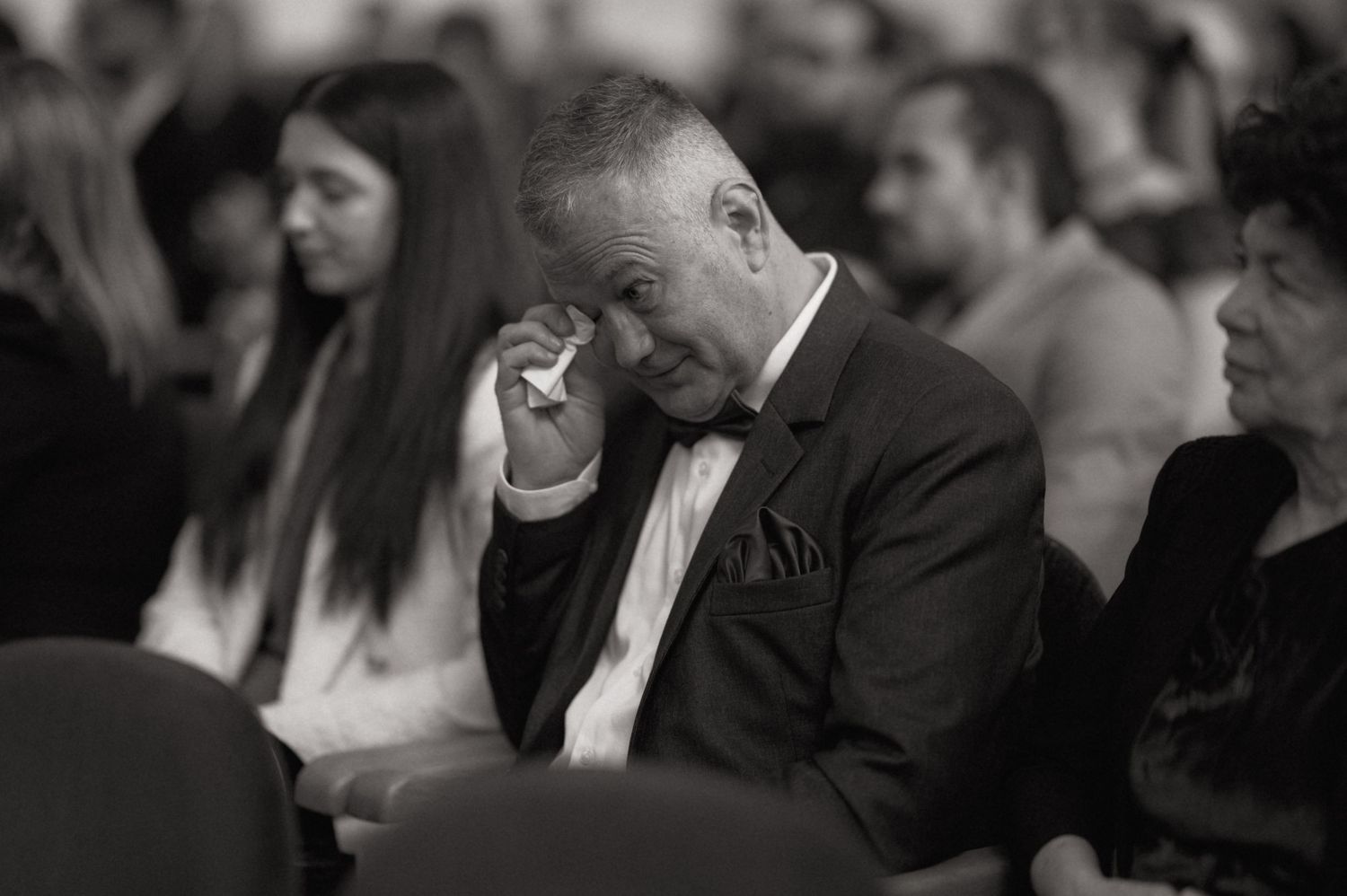 Black and white photo of an emotional man in a tuxedo wiping tears at a formal event.