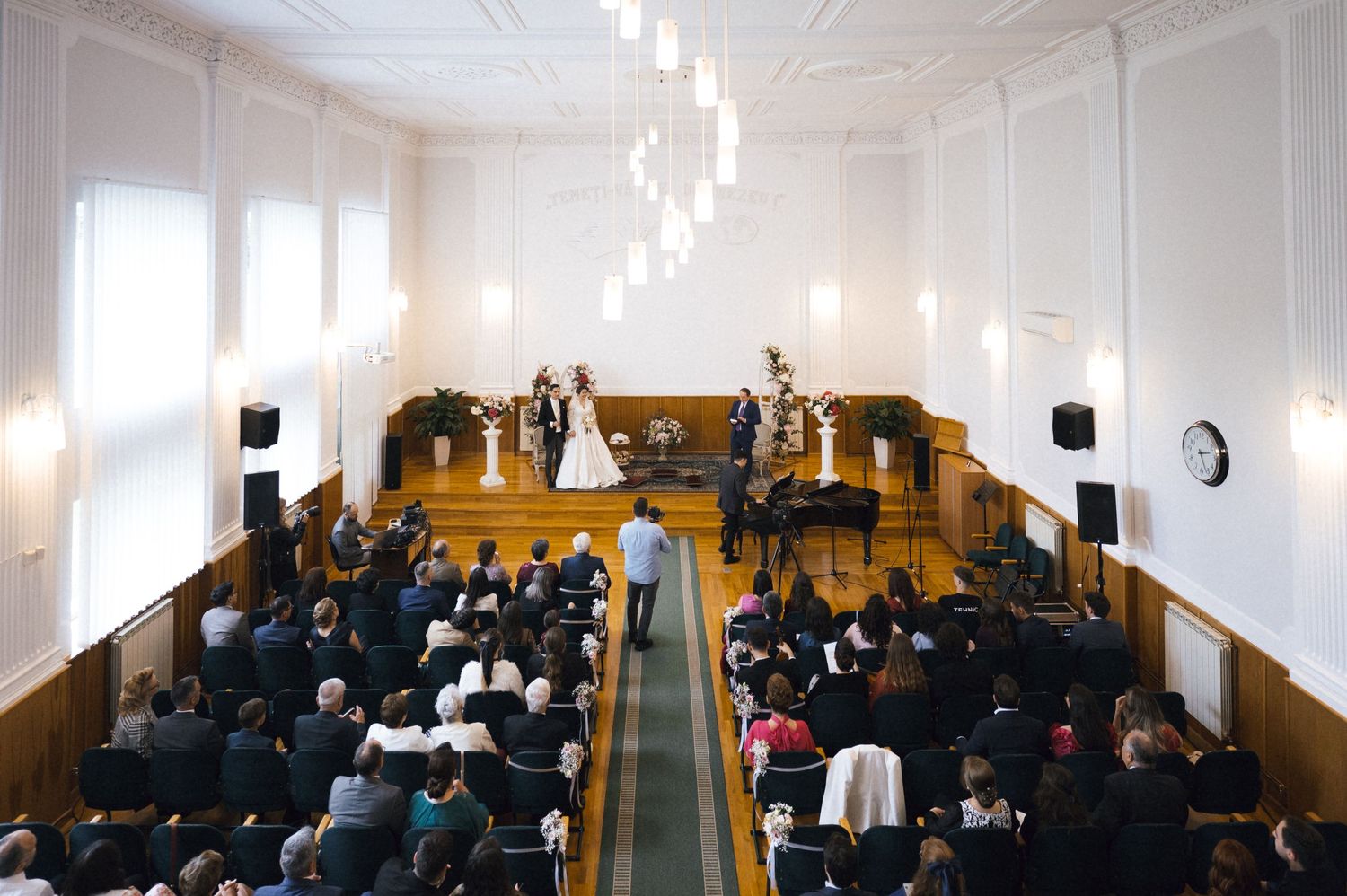 Elegant wedding ceremony in a grand white hall with chandeliers, guests seated as couple stands at the altar.
