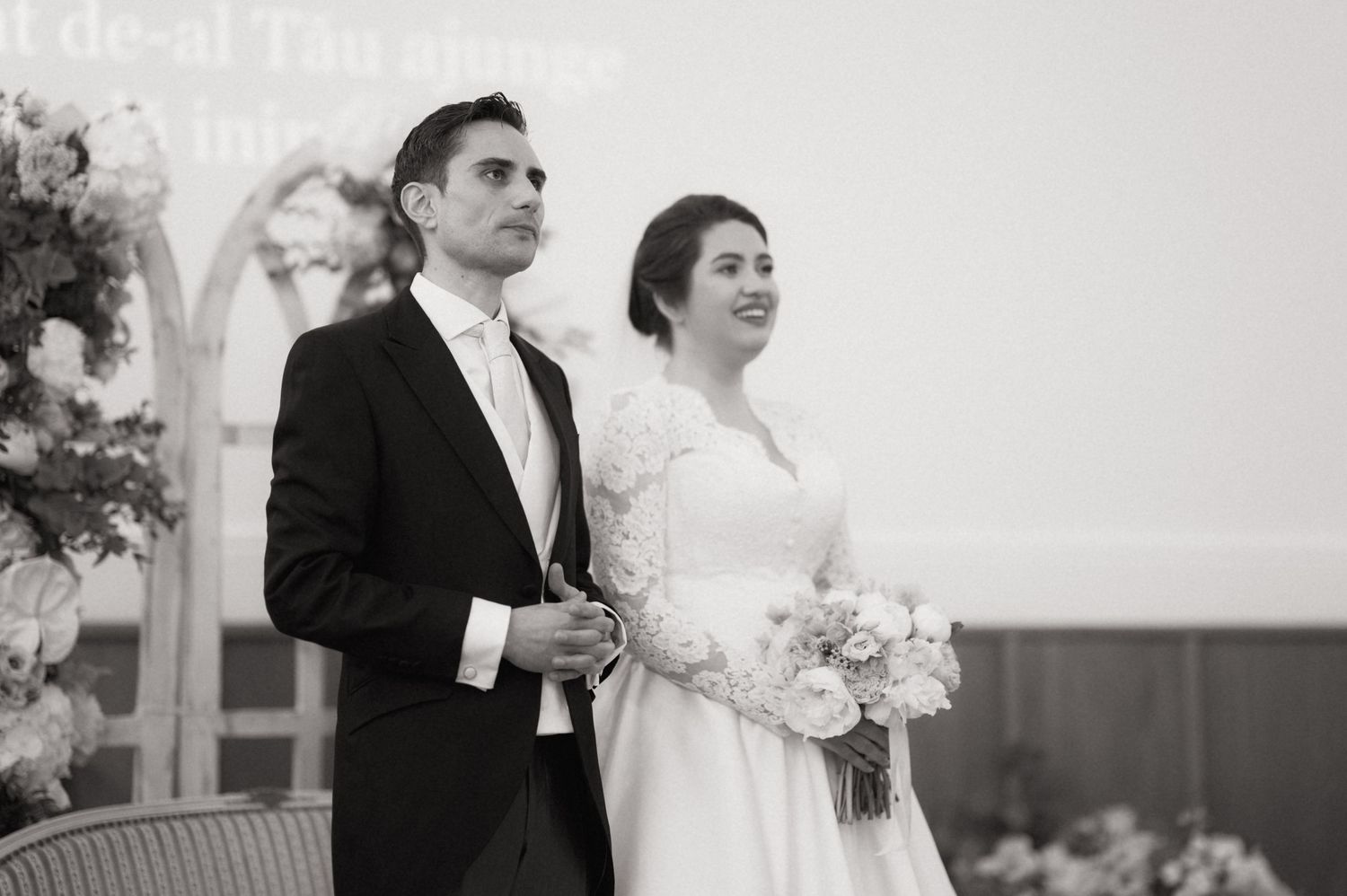 Black and white wedding photo of bride holding bouquet and groom in tuxedo standing together at altar ceremony.