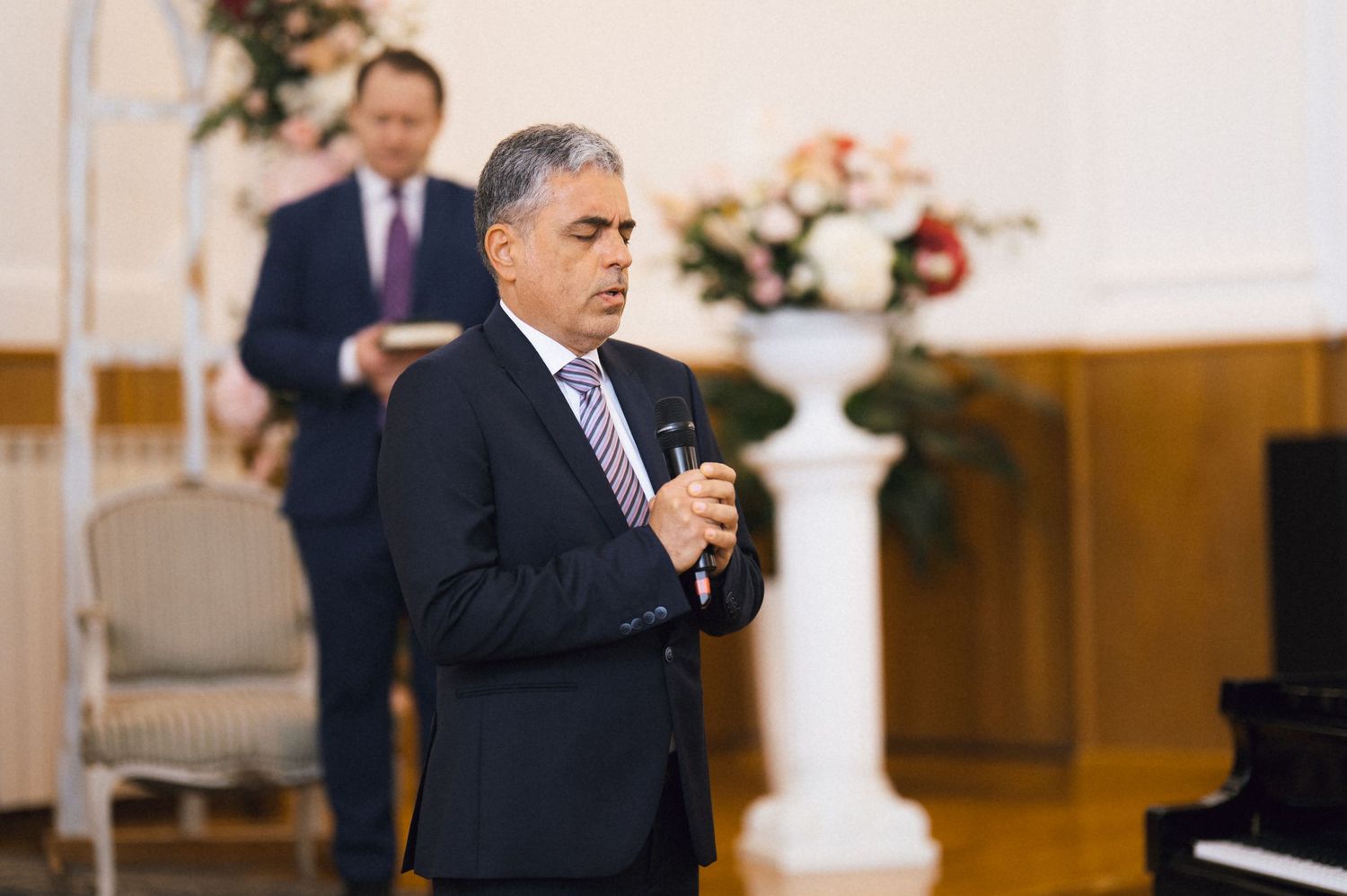 Man in navy suit speaking into microphone at wedding ceremony with floral arrangements and pianist in background.