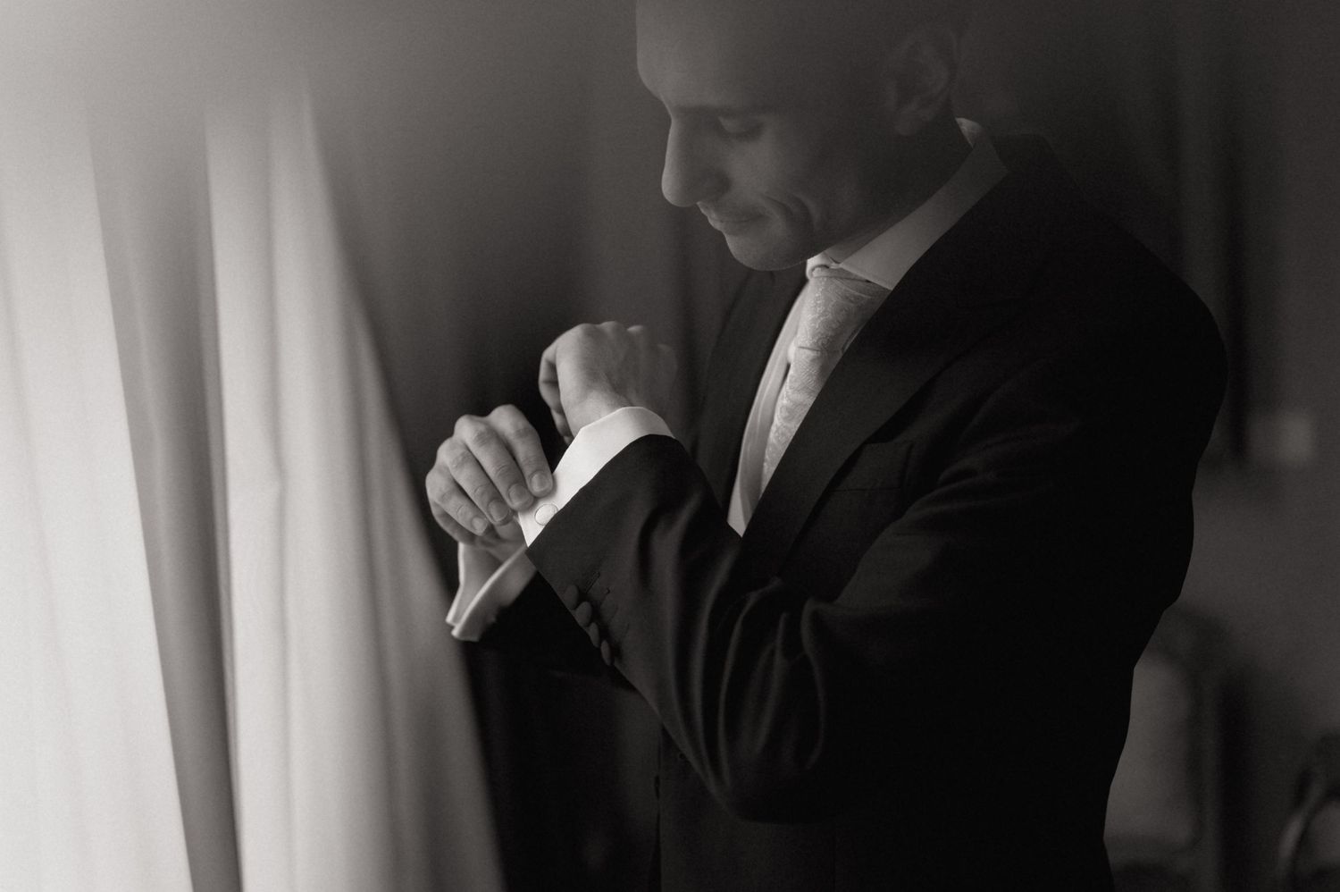 Black and white photo of an elegant groom in a suit adjusting his cufflinks near a window.