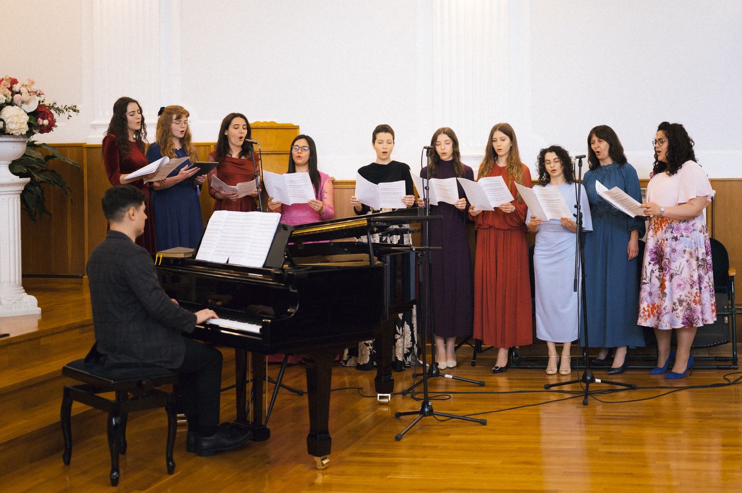 A choir of young women performs on stage, accompanied by a pianist playing a grand piano in a concert hall.