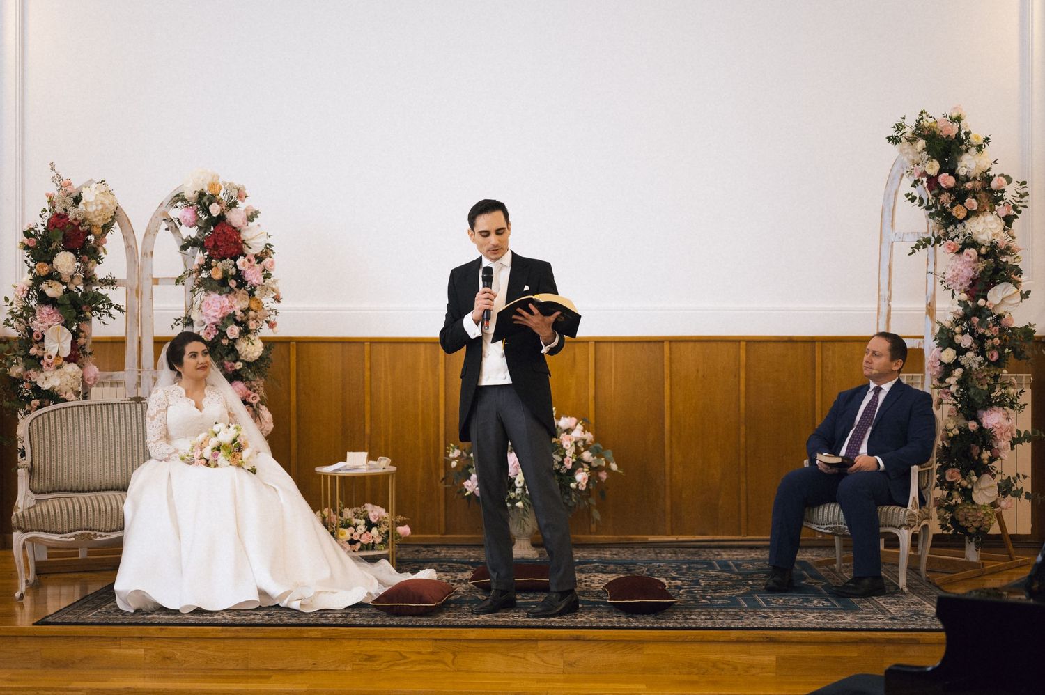 Officiant reads from book during wedding ceremony with bride in white gown and seated guest beside floral arrangements.