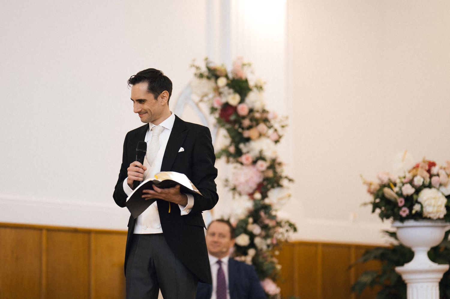 Man in tuxedo giving a speech at a wedding ceremony, holding a book, with floral arrangements in the background.