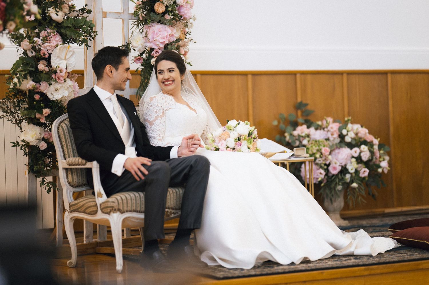 Bride and groom sitting together at wedding ceremony, surrounded by pink floral arrangements in elegant venue.