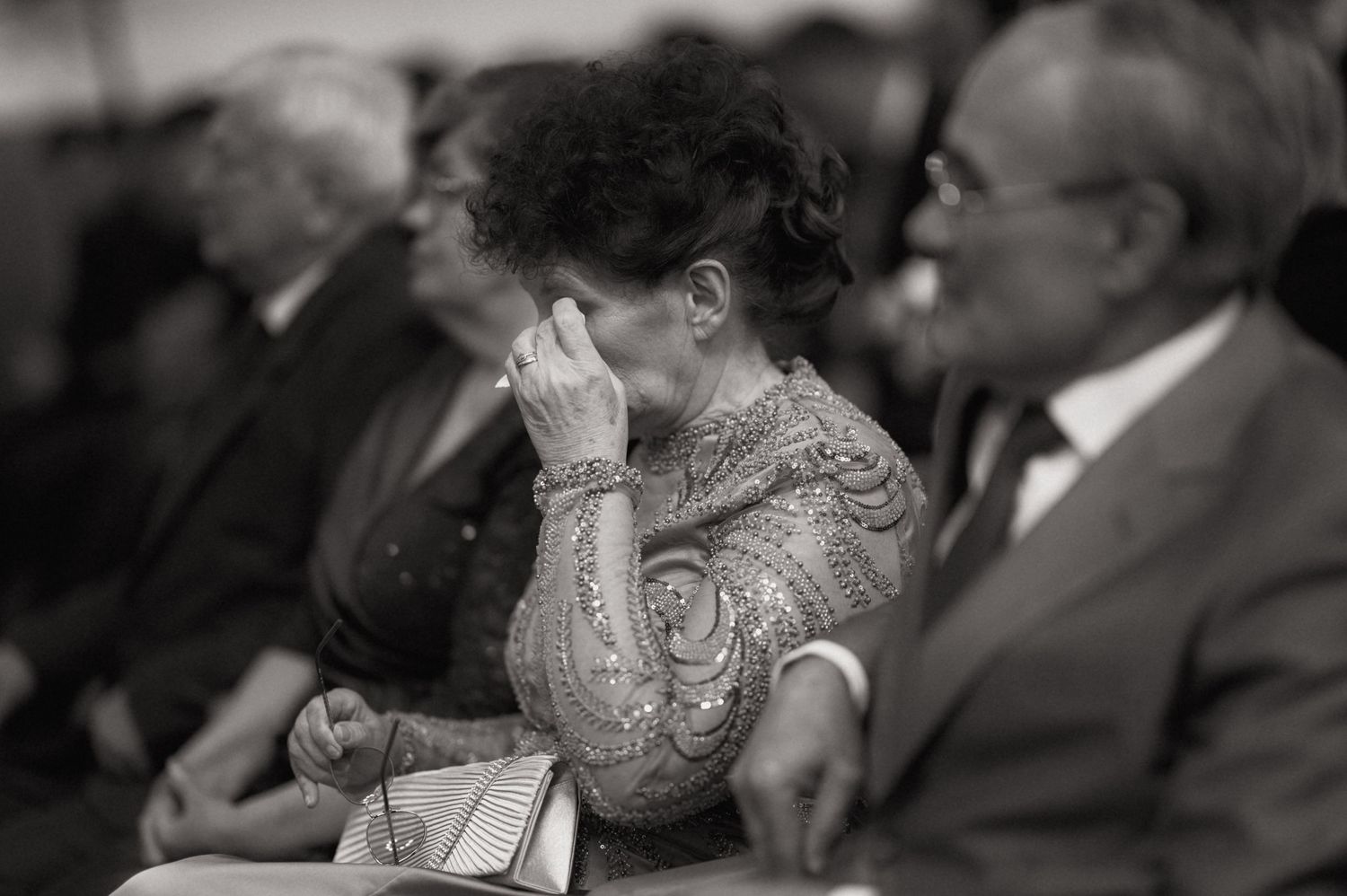 Black and white photo of an emotional elderly woman covering her face at a formal gathering.