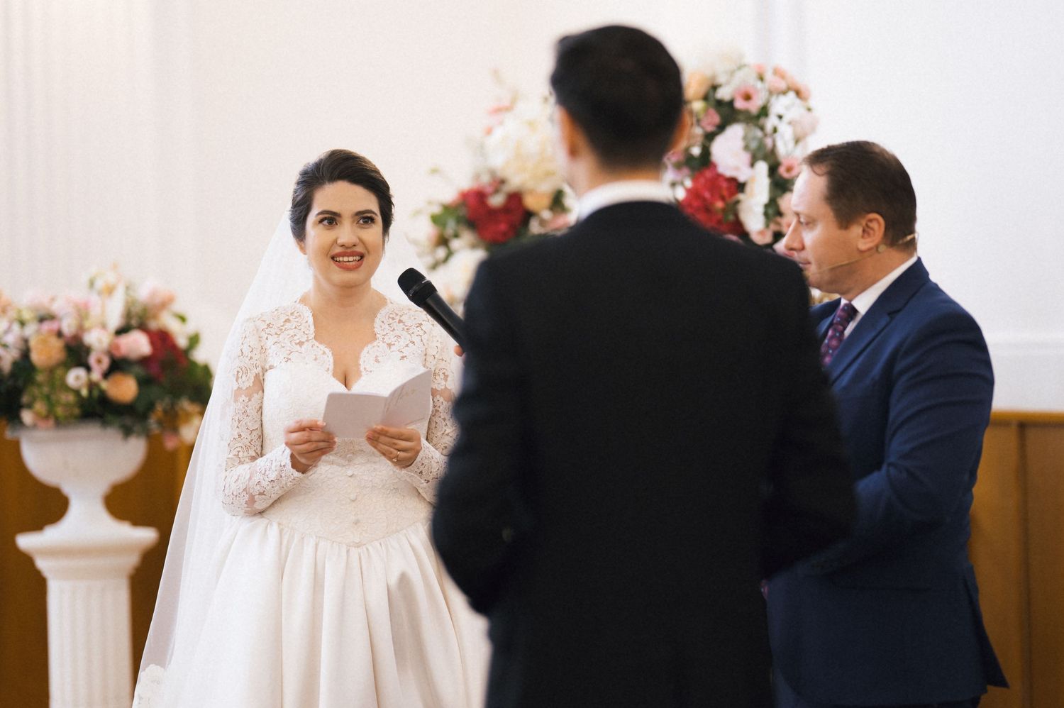 Bride in white lace gown reads vows at wedding ceremony altar with groom and officiant, floral arrangements in background.