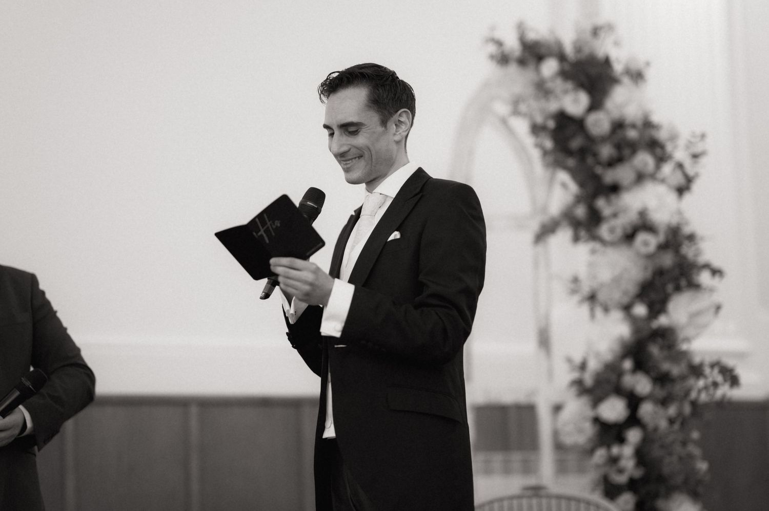 Man in tuxedo smiling while reading from a book at a formal wedding reception in black and white.