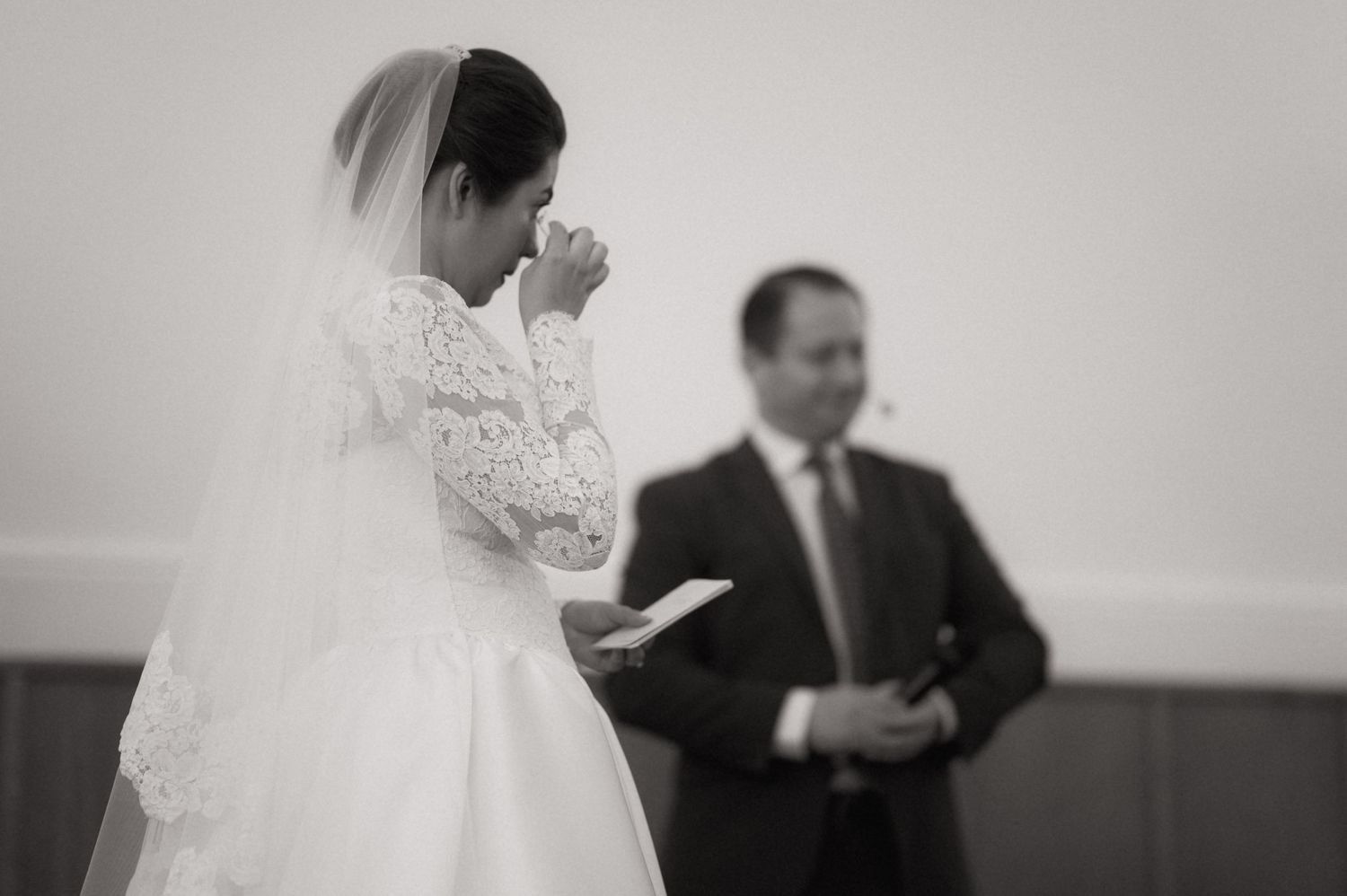 Black and white wedding photo of a bride in lace gown wiping tears while groom stands in background.