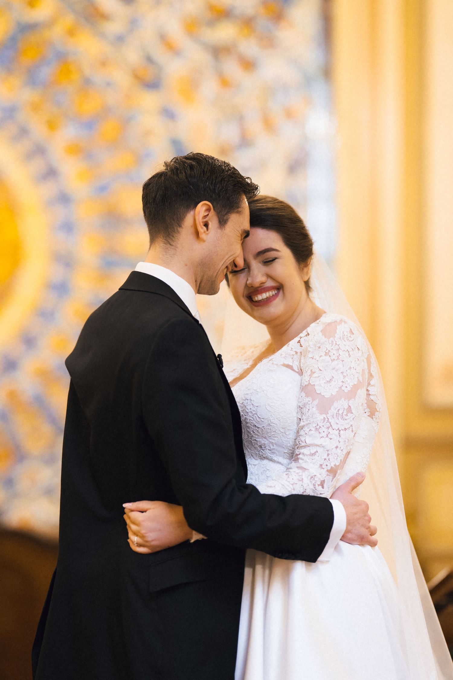 Smiling bride in lace wedding dress embraces groom in black suit during romantic wedding ceremony.
