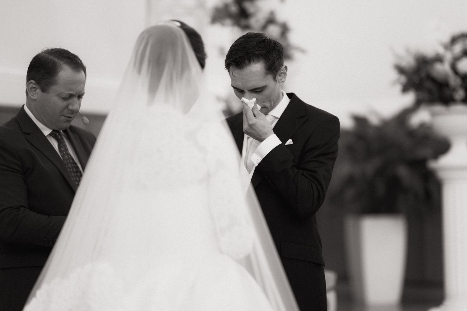 Emotional groom wiping tears during wedding ceremony in black and white photo with bride in veil.