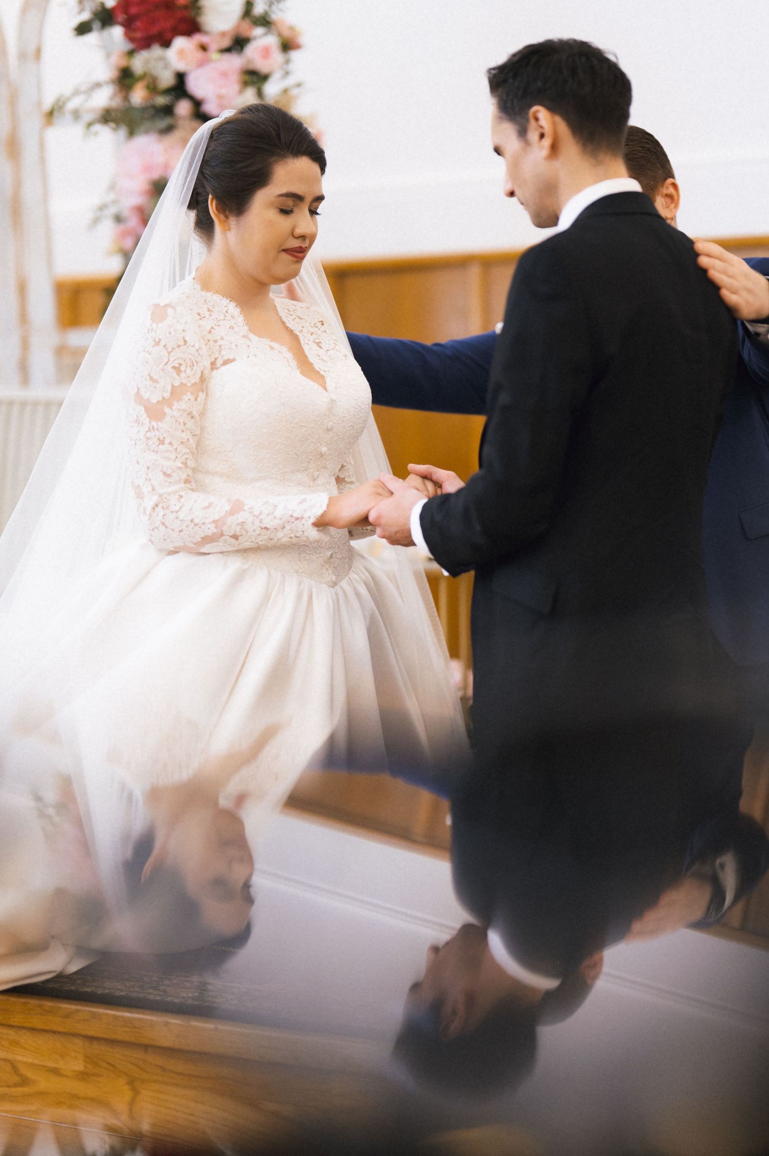 Bride in lace wedding gown and veil holds hands with groom in black suit during church wedding ceremony.