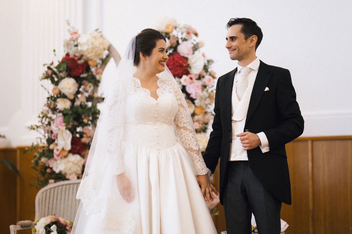 Bride and groom smiling at each other holding hands at wedding ceremony with floral backdrop.