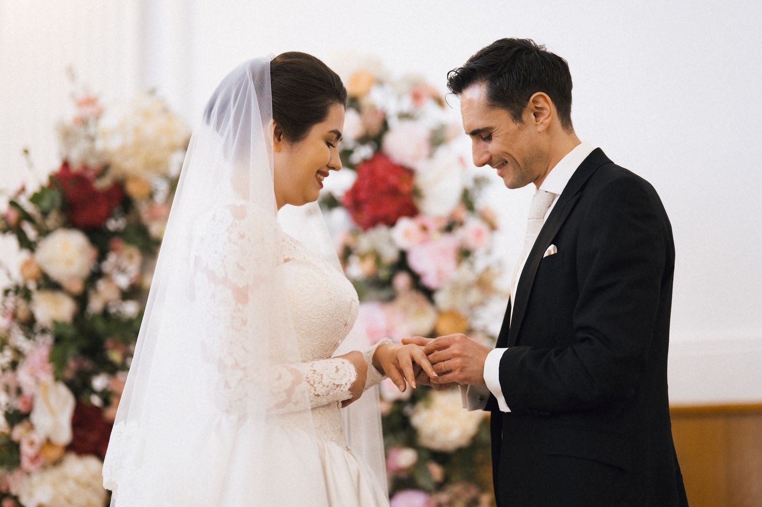 Groom placing wedding ring on bride's finger at altar with floral arrangement backdrop.
