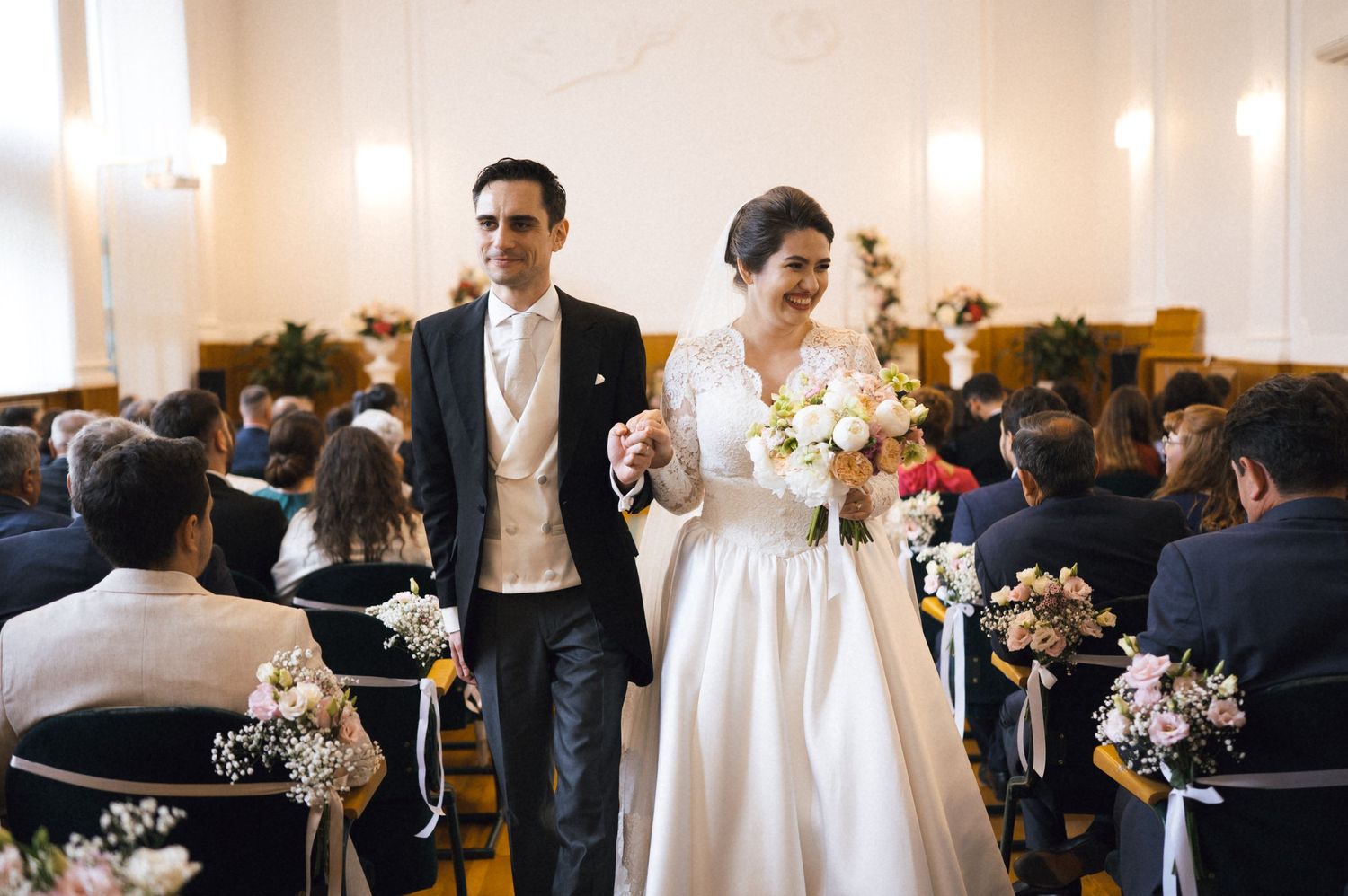 Bride and groom walking down the aisle together, smiling, as guests look on in an elegant wedding ceremony hall.