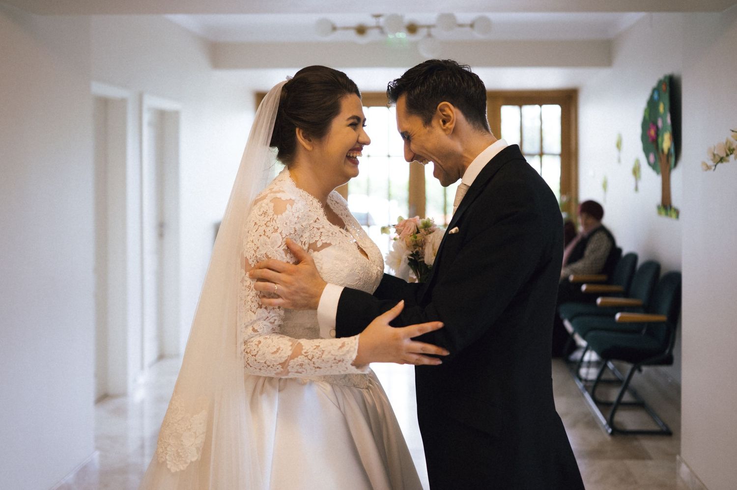 Bride and groom share a joyful embrace, smiling at each other in a bright hallway on their wedding day.