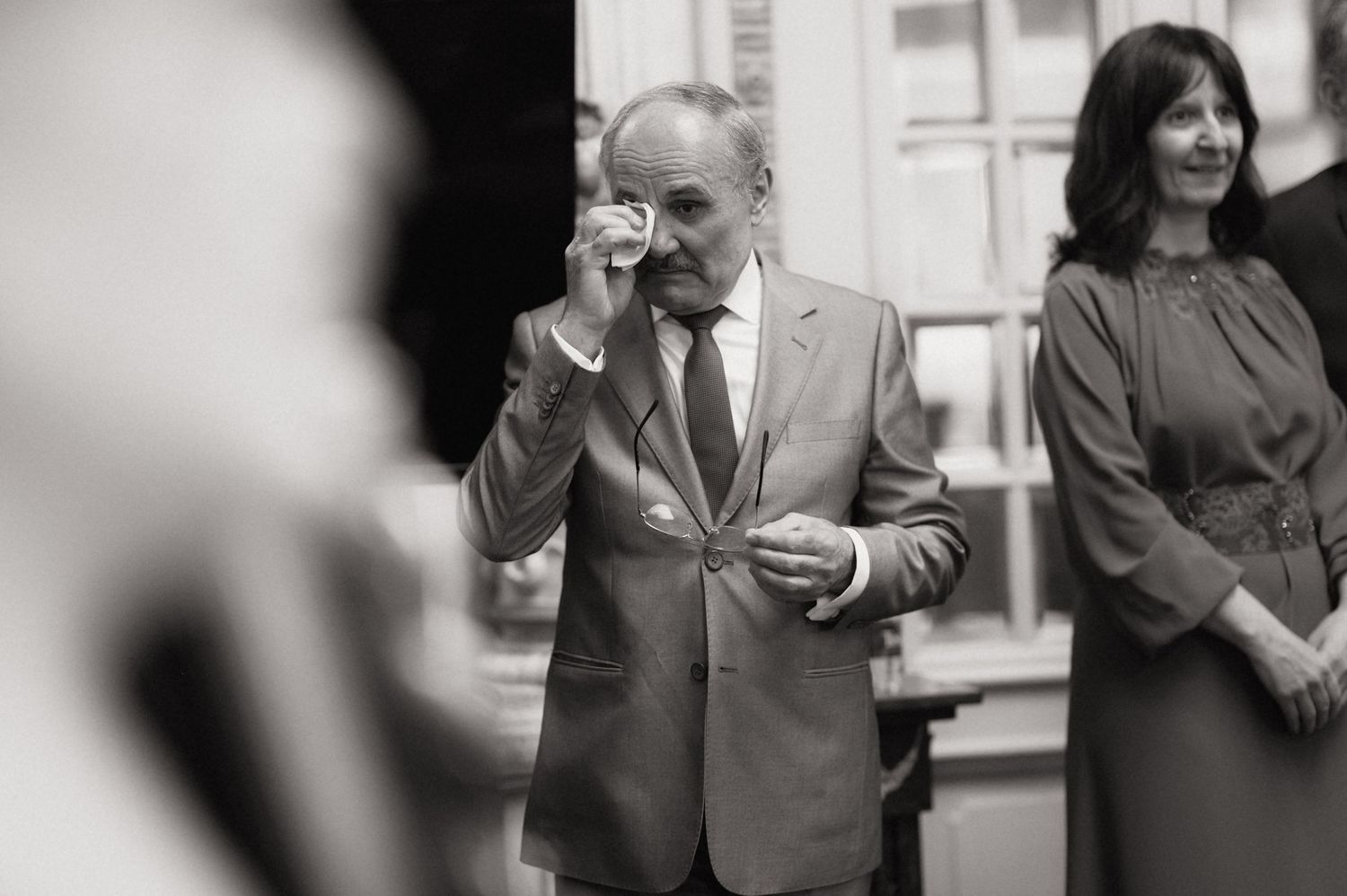 Black and white photo of an emotional man in a suit wiping his eye at a formal event, woman smiling behind him.
